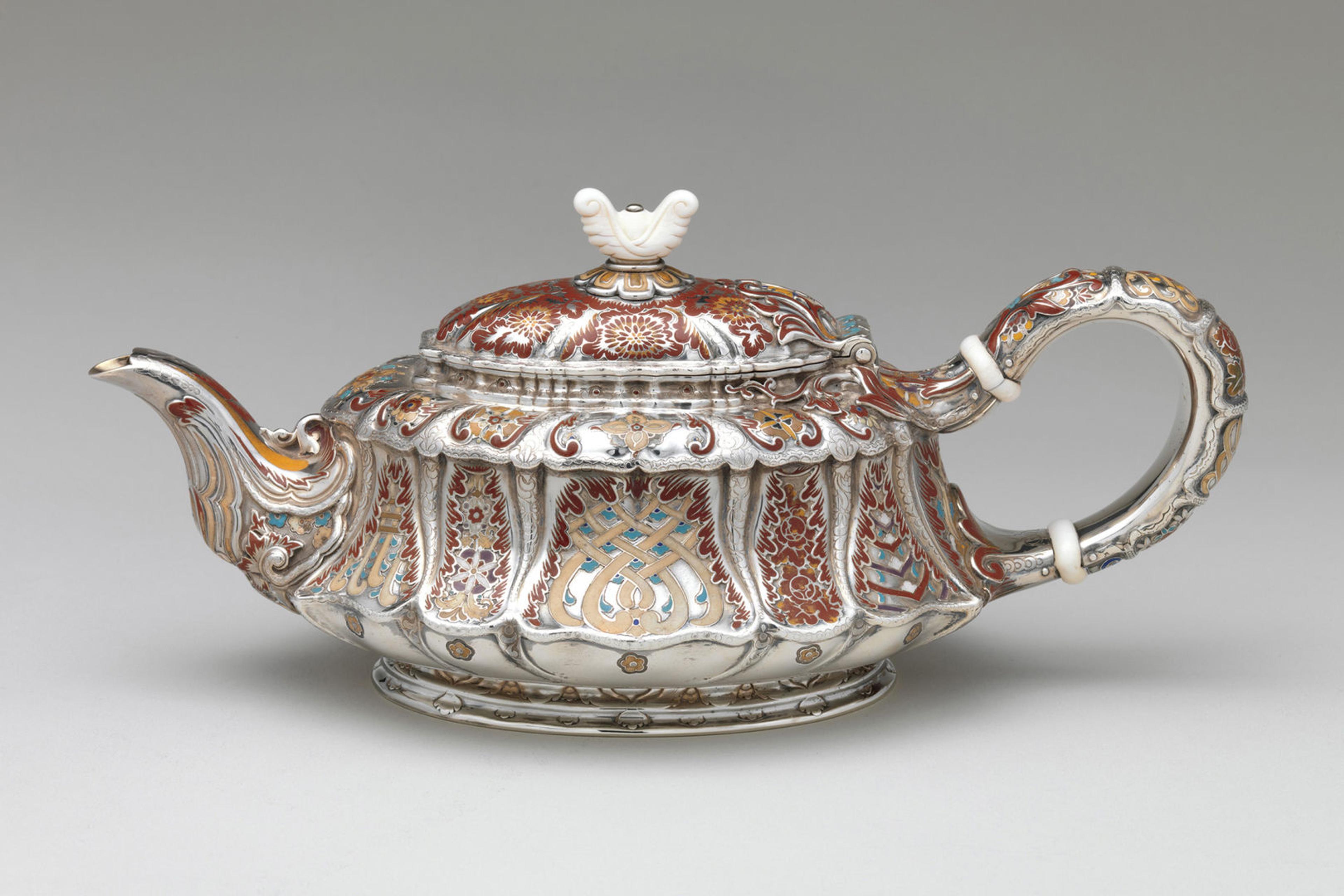 A short but elongated silver teapot with an ivory lid finial adorned with abstract floral motifs rendered in maroon, orange, taupe, and turquoise enamel.