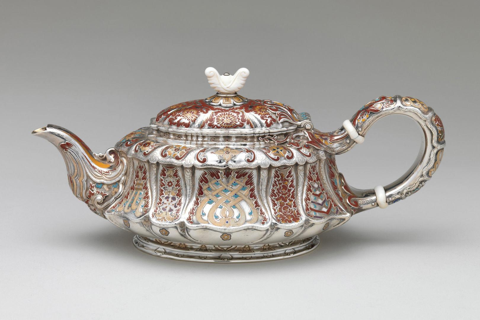 A short but elongated silver teapot with an ivory lid finial adorned with abstract floral motifs rendered in maroon, orange, taupe, and turquoise enamel.