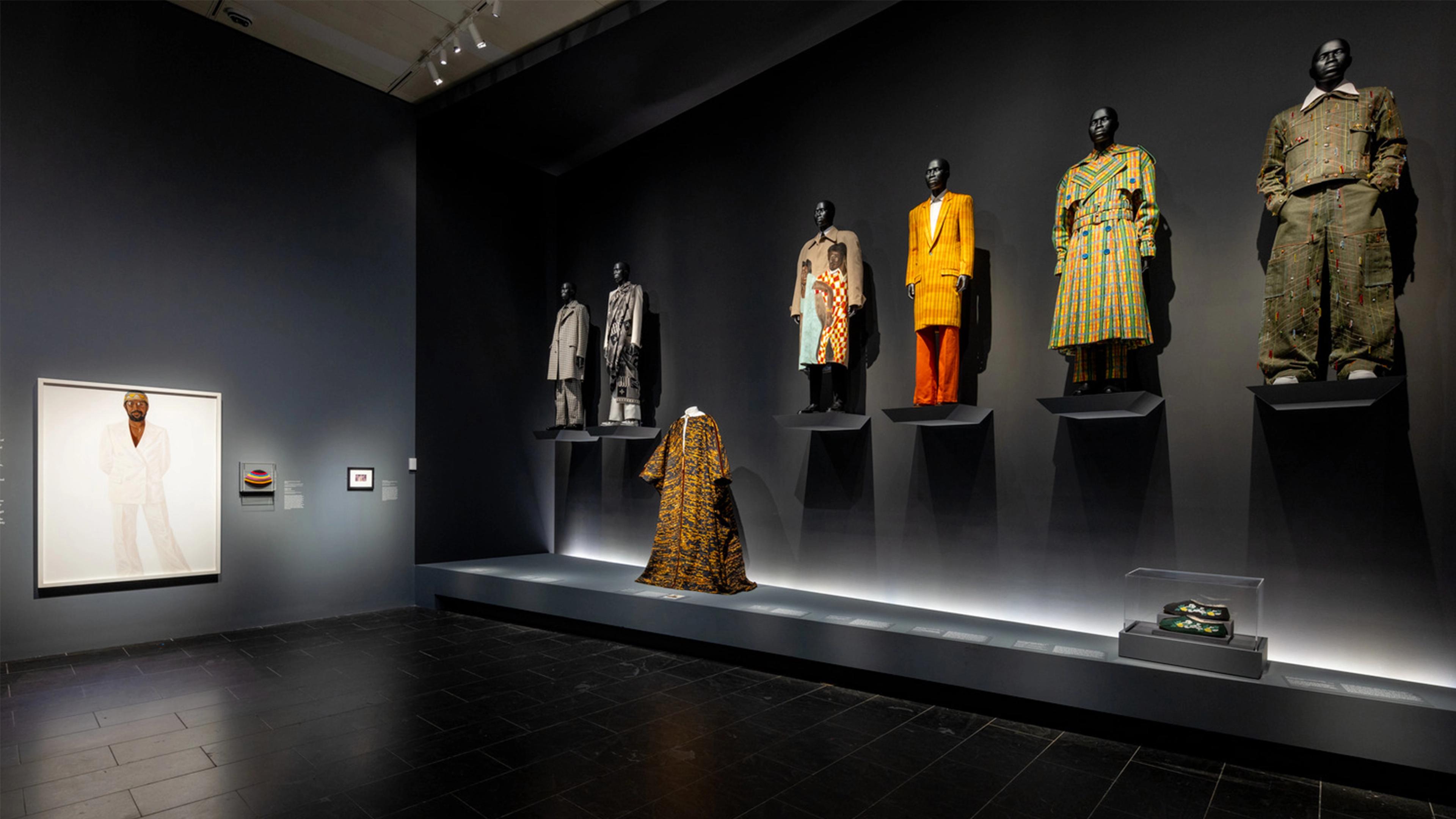 A dimly lit fashion exhibit displays mannequins in vibrant, patterned suits on platforms, with a large portrait and artifacts on a dark wall, creating a modern and elegant ambiance.