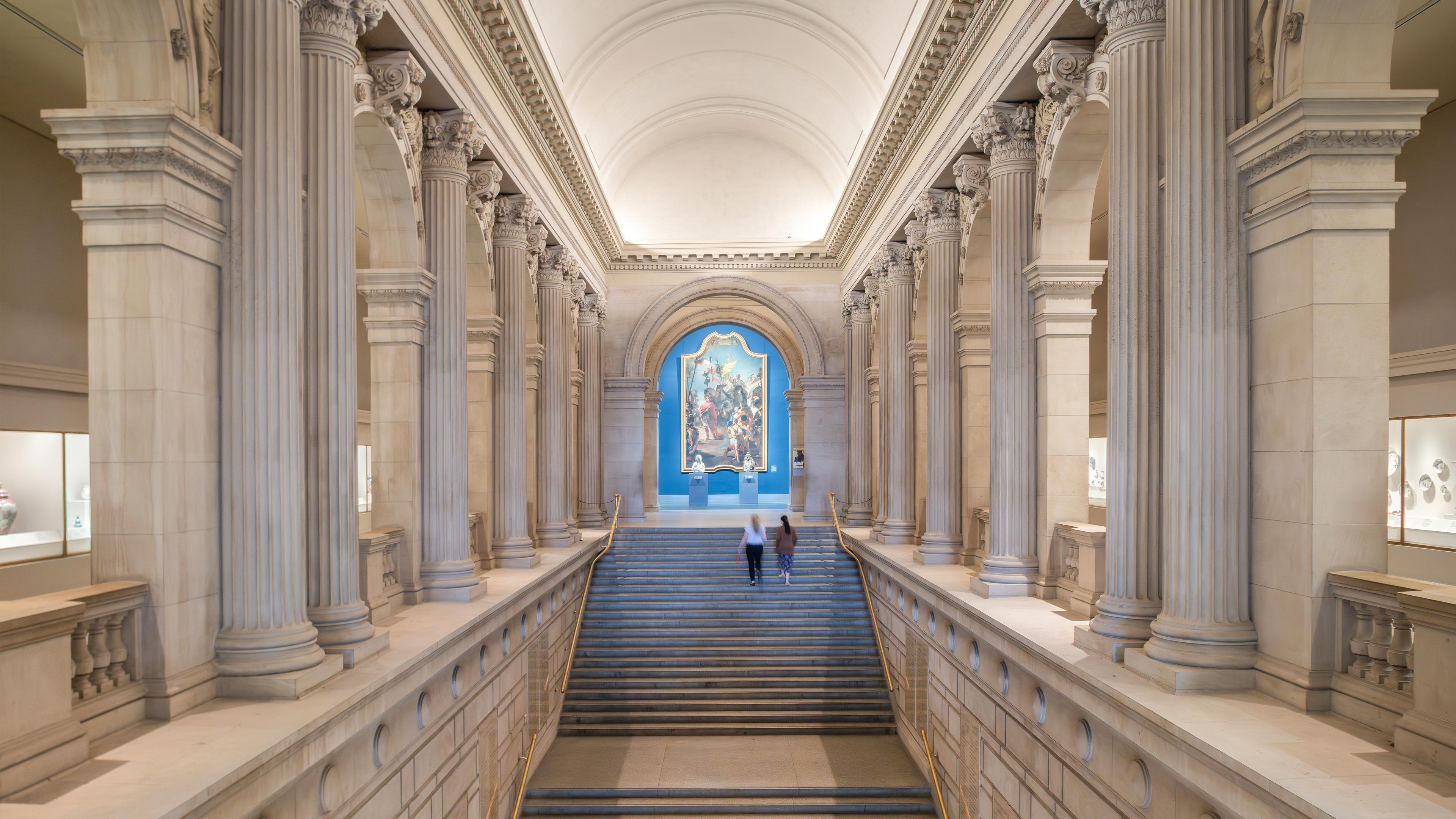 A grand museum interior features light tall marble columns, a wide set of stairs with a large painting at the top, and two visitors walking up the steps.