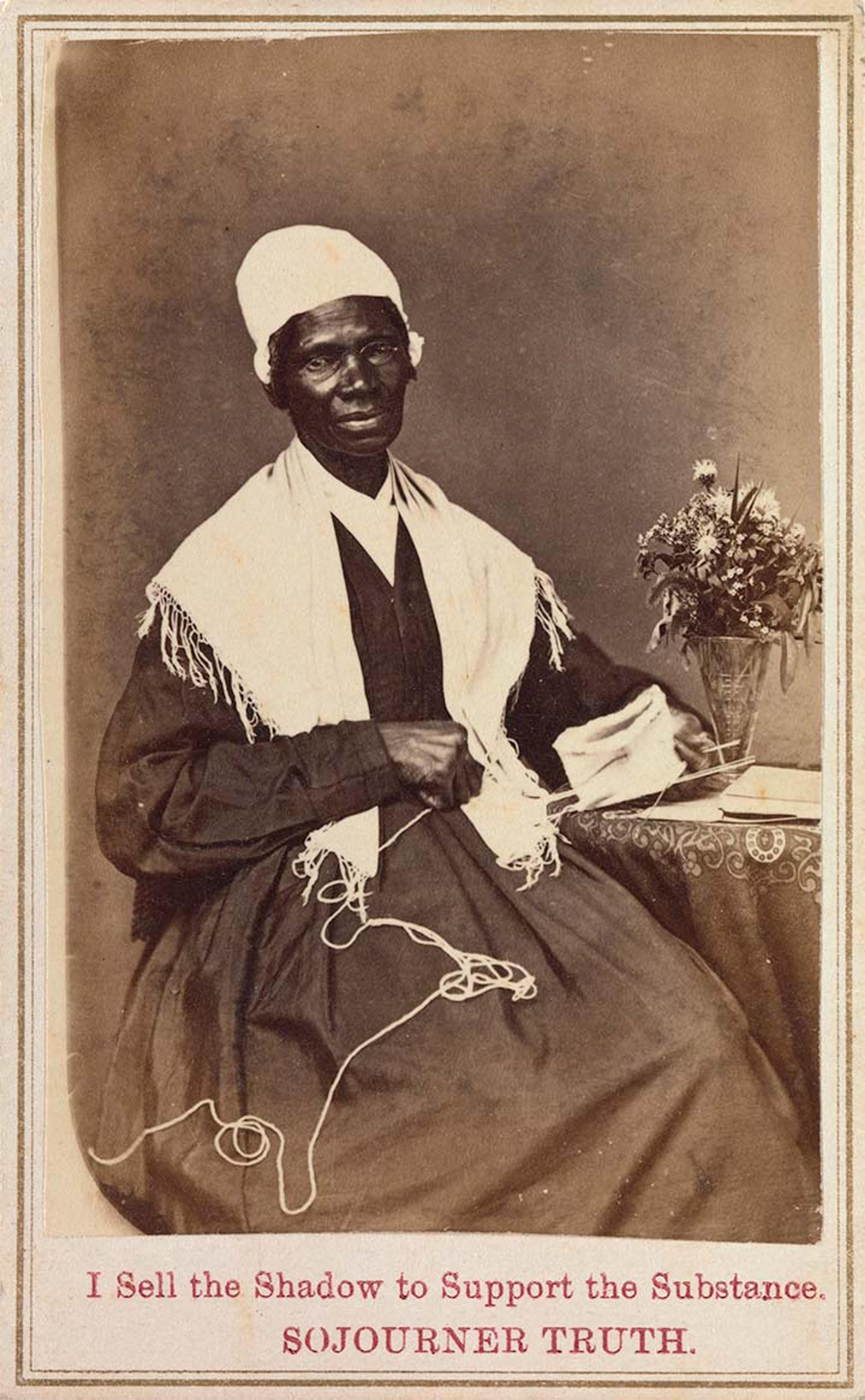 Sojourner Truth stops knitting to look at the camera. She sits upright in a chair resting her left arm on a table. On the table is also a vase of flowers.