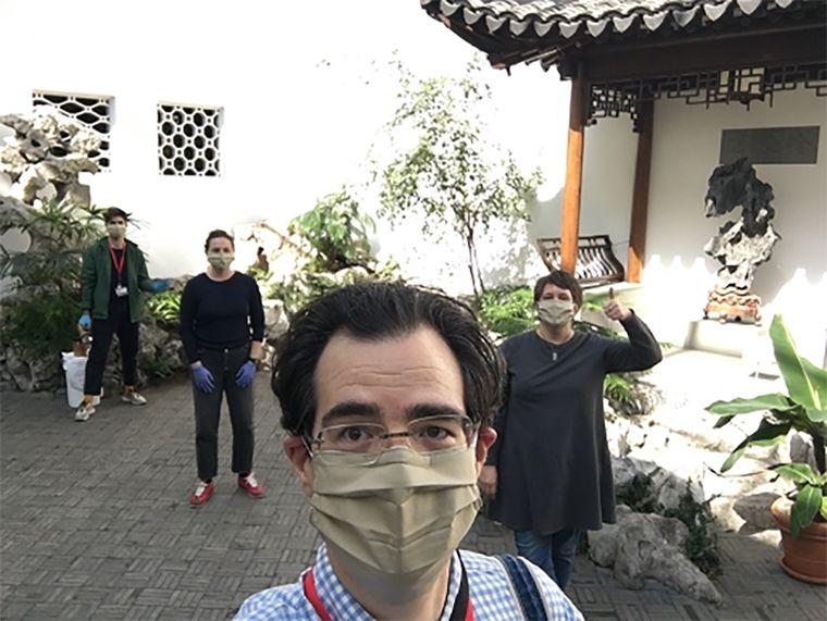 A Met employee in a facemask faces the camera in The Met's interior Chinese Garden Court. Three other people wearing facemasks stand behind him and wave.