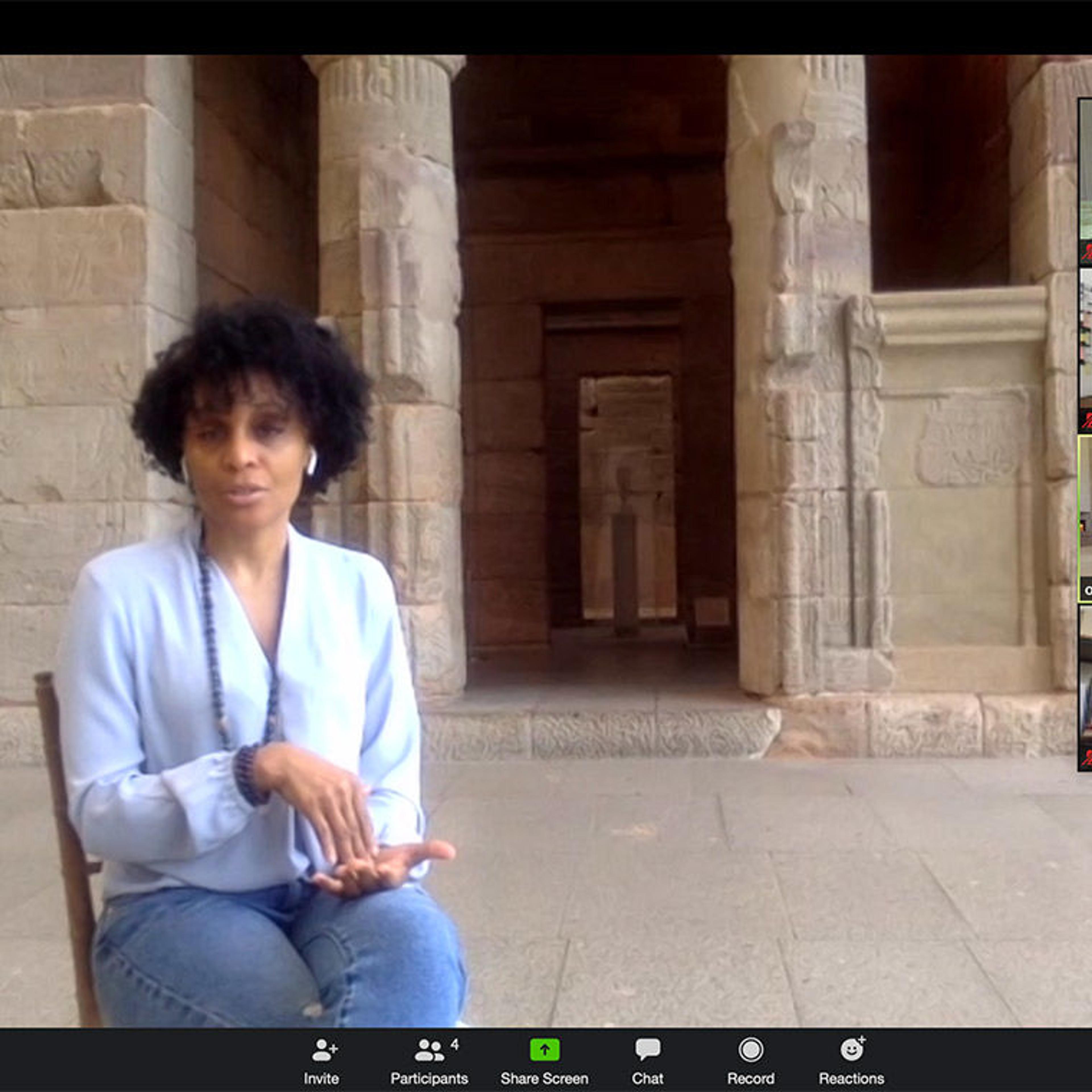 A zoom screen capture of a seated woman in front of an ancient temple at The Met being interviewed by a production crew.