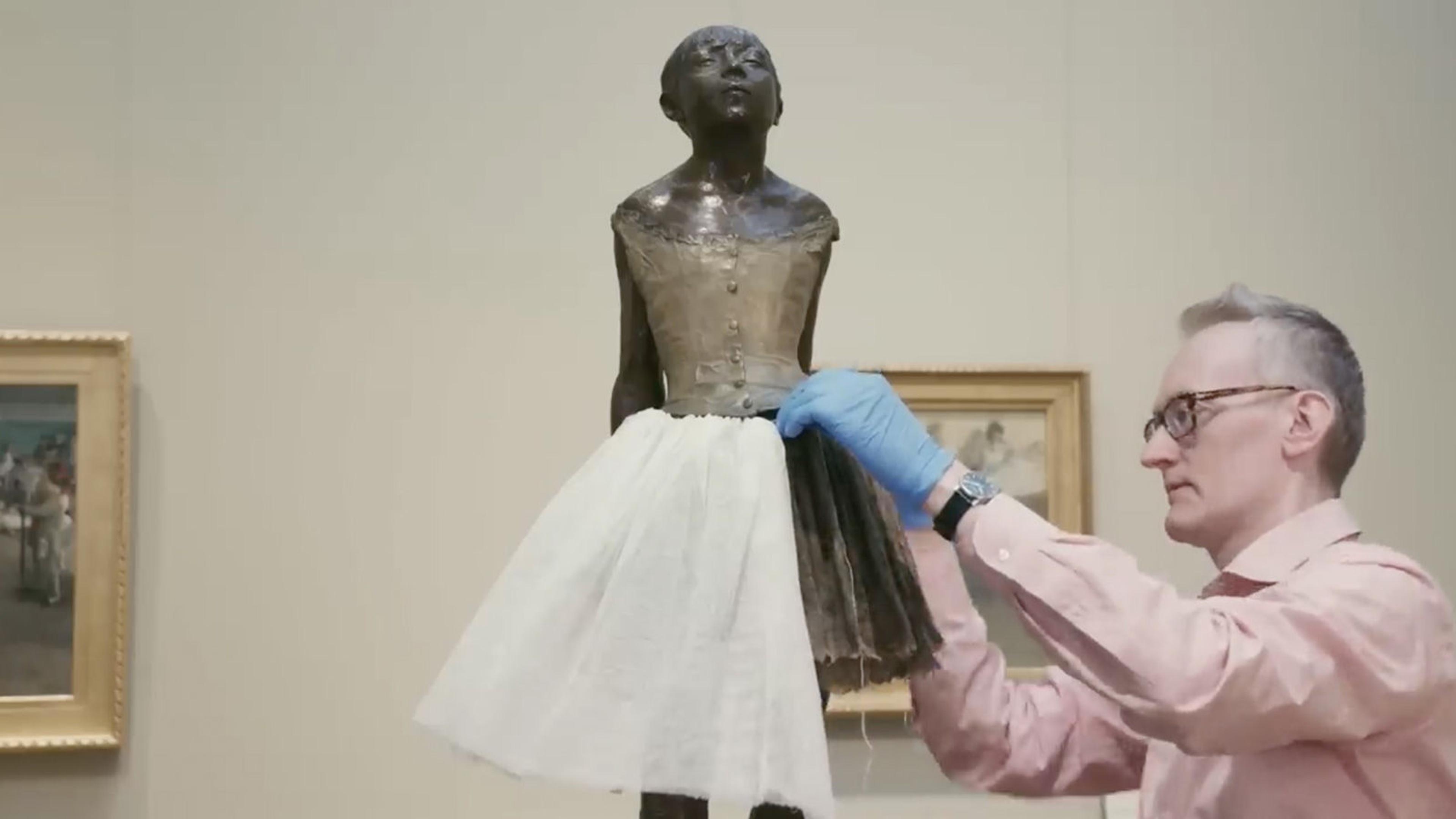 A conservationist in a gallery setting stitches the tutu on Edgar Degas's sculpture, The Little Fourteen-Year-Old Dancer.
