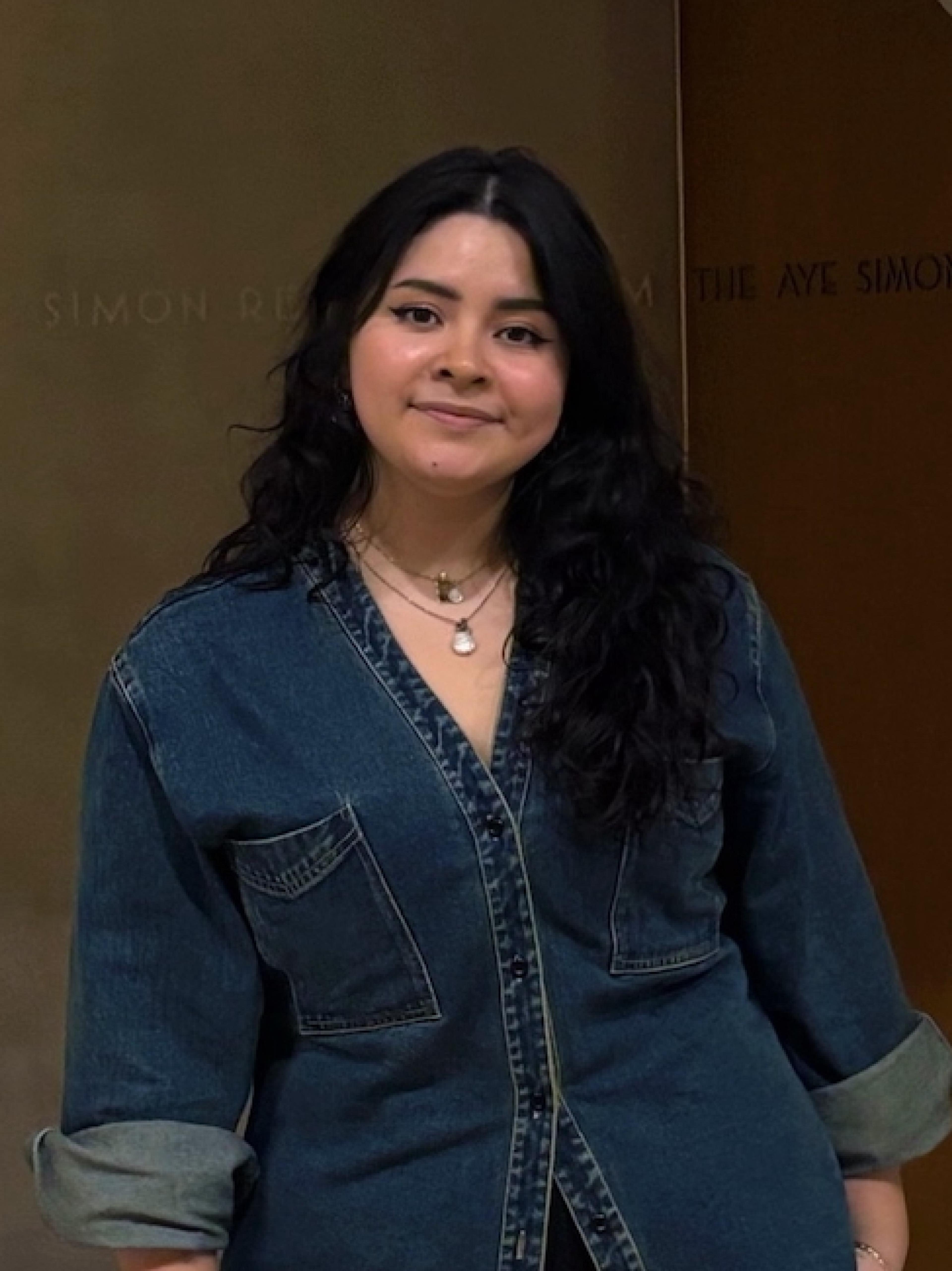 Photo of Daniela Hernandez wearing a dark, long sleeve, jean dress. Dani is looking at the camera. 