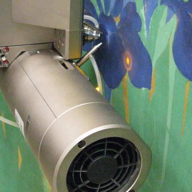 Photographed up-close, from three-quarter view, a piece of silver scientific equipment shines a precise bulls-eye light on a painting of blue irises.