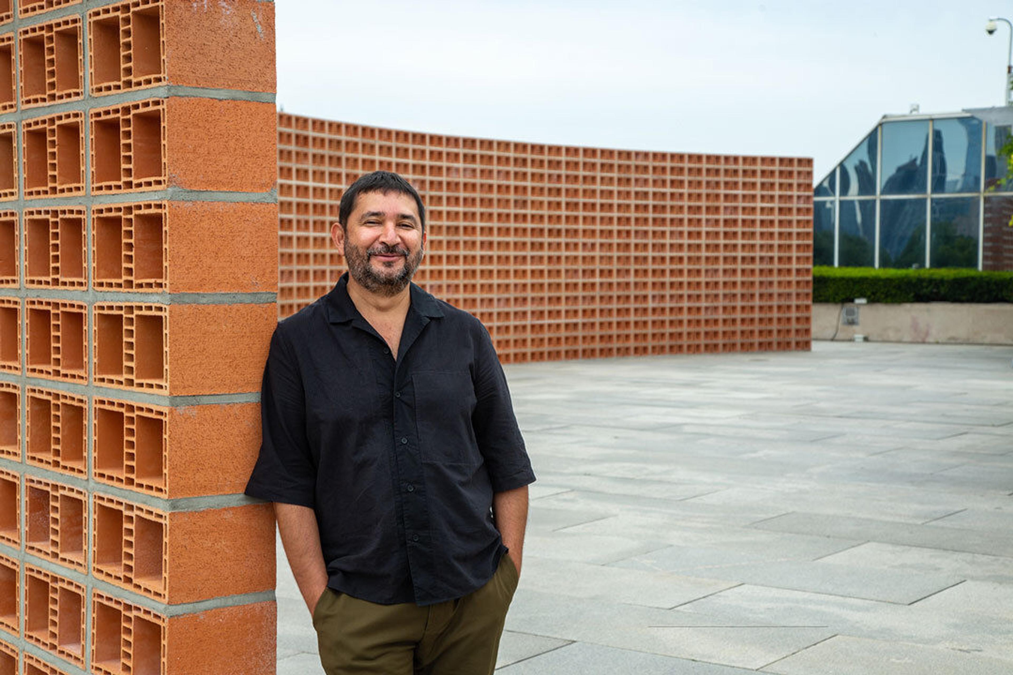 Man standing in a black shirt and green pants standing up again a brick wall.