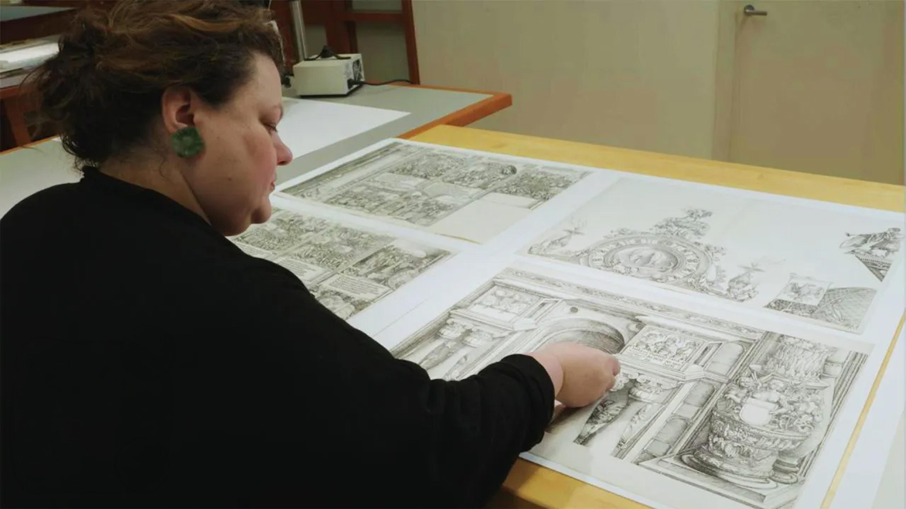A conservator in a black sweater sits at a table and works on a panel of a larger woodblock print.