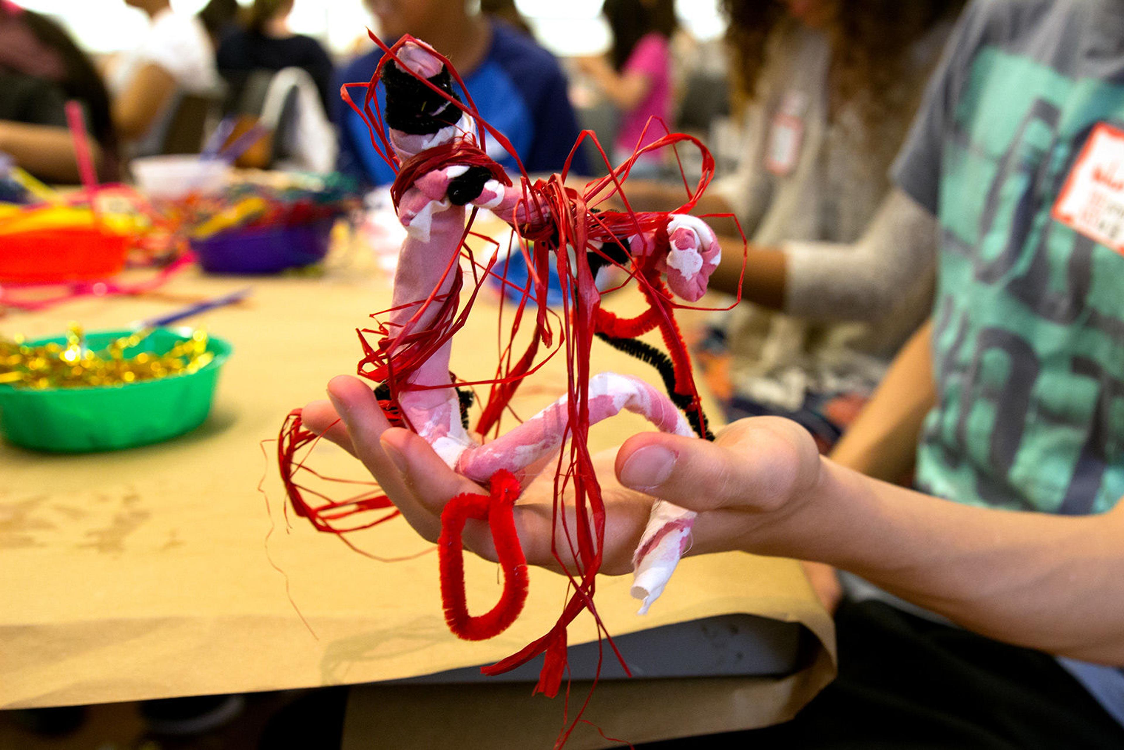 Kid holds up mobile he is working on made out of pink and red pipe cleaners, twist tyes, and dyed cloth.