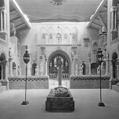 Medieval-style hall with brick arches and statues, illuminated by lanterns arranged around a central stone structure.