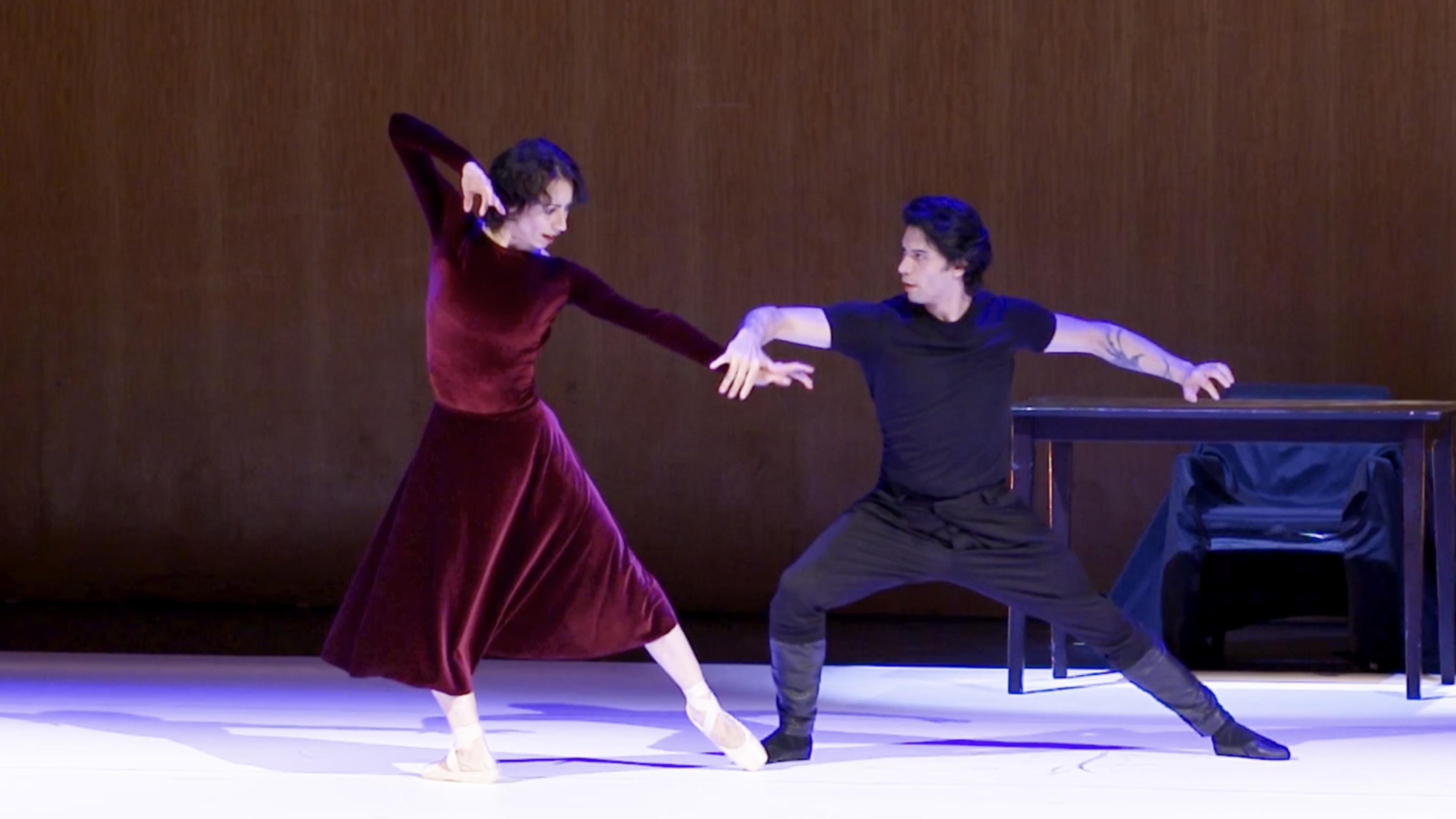 Two dancers in performance with dramatic poses. The woman at left wears a red velvet dress, while the man at right is clad in a black shirt and pants.