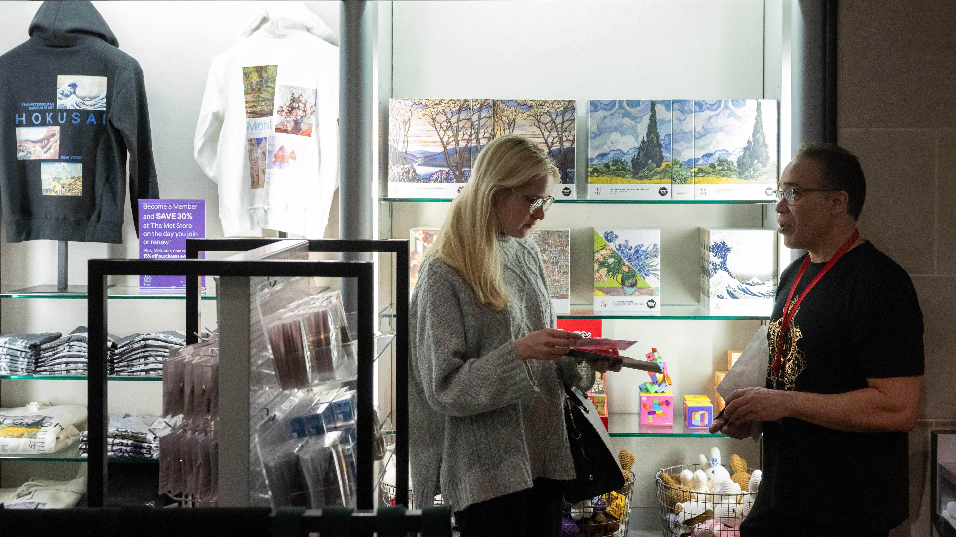 A woman holds products from The Met Store while speaking with a staff member. Art-inspired hoodies, puzzles, and children's toys can be seen behind them.