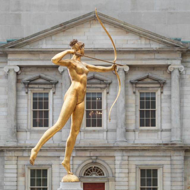 Gold statue of a nude woman, pointing a bow and arrow in the direction of a stately gray facade.