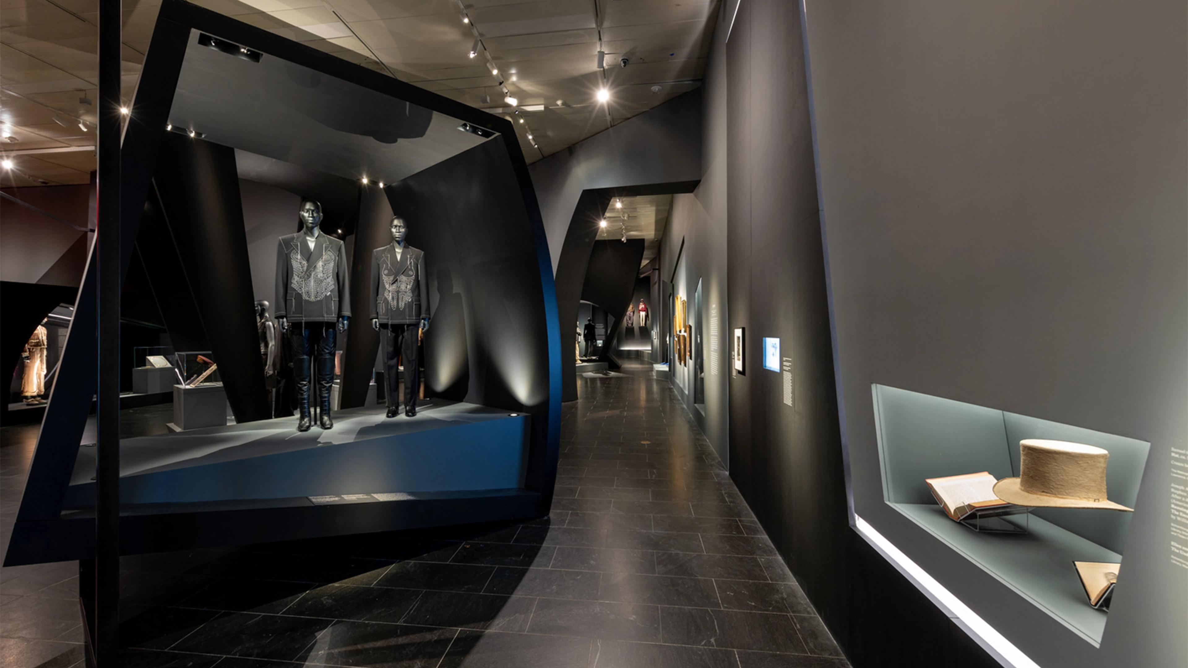 Sleek museum exhibit with black walls, spotlighting mannequins in embellished suits. Nearby, a display case features an open book and a wide-brimmed hat. Modern and sophisticated ambiance.