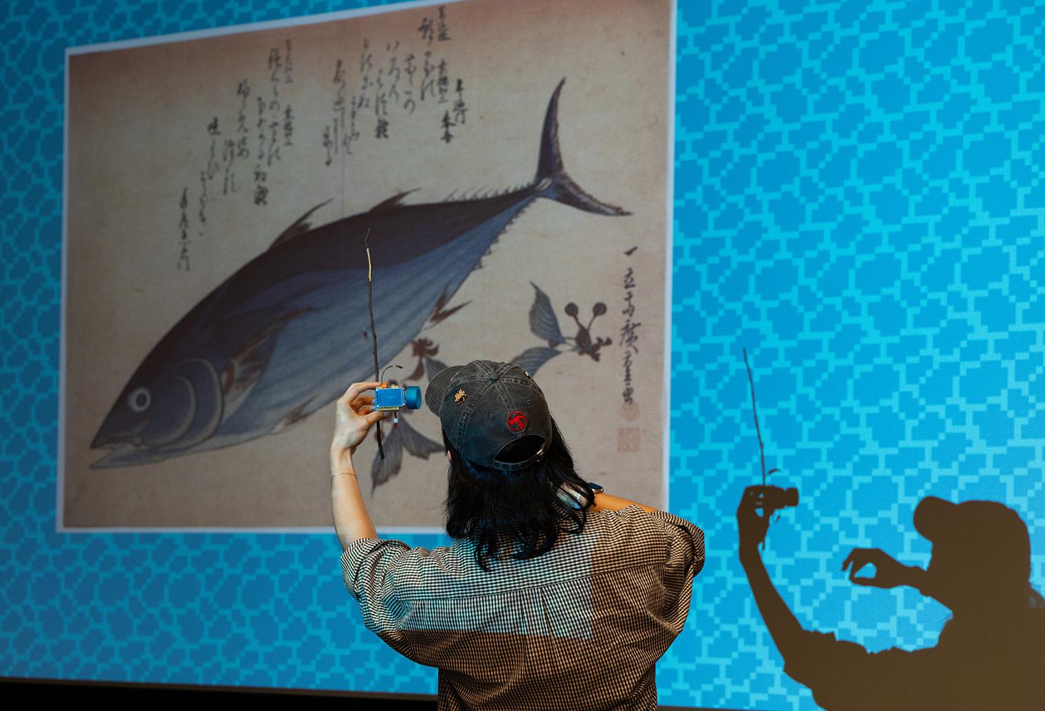 A person in a cap uses a smartphone to take a photo of a large fish illustration projected on a patterned blue wall. Their silhouette is visible.