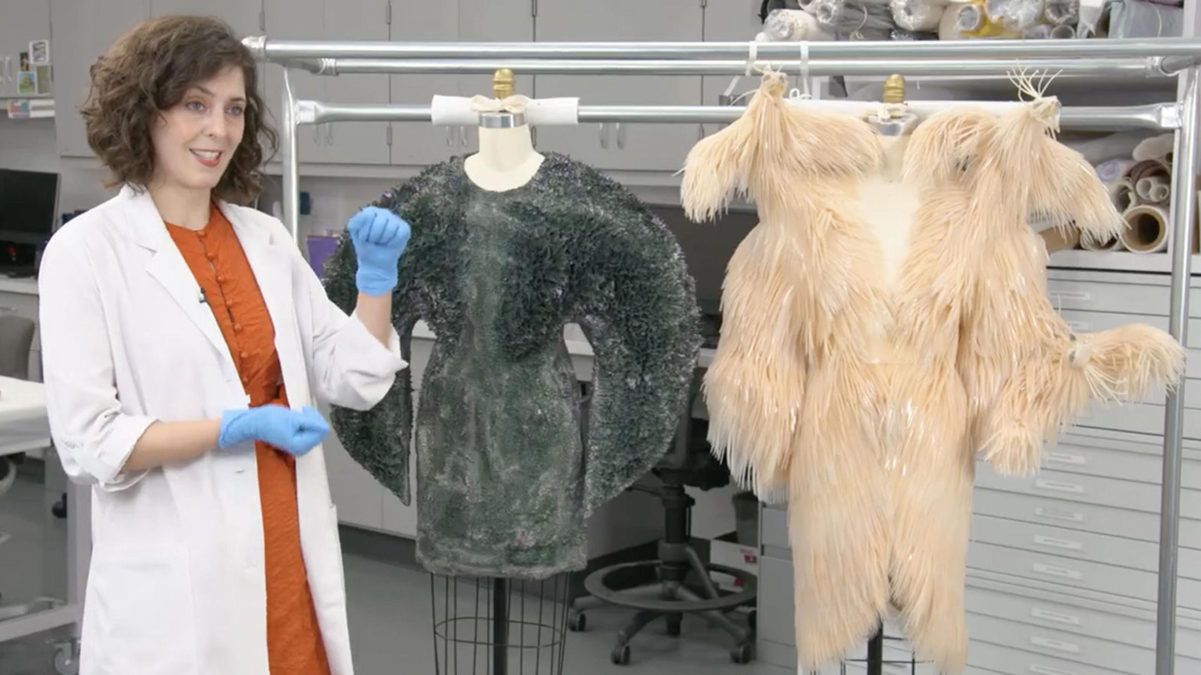 A conservationist in a white gown and blue gloves gestures towards two hanging costumes to her left: the first is a greenish-blue dress, and the second is a peach color. Both are made with unconventional materials.