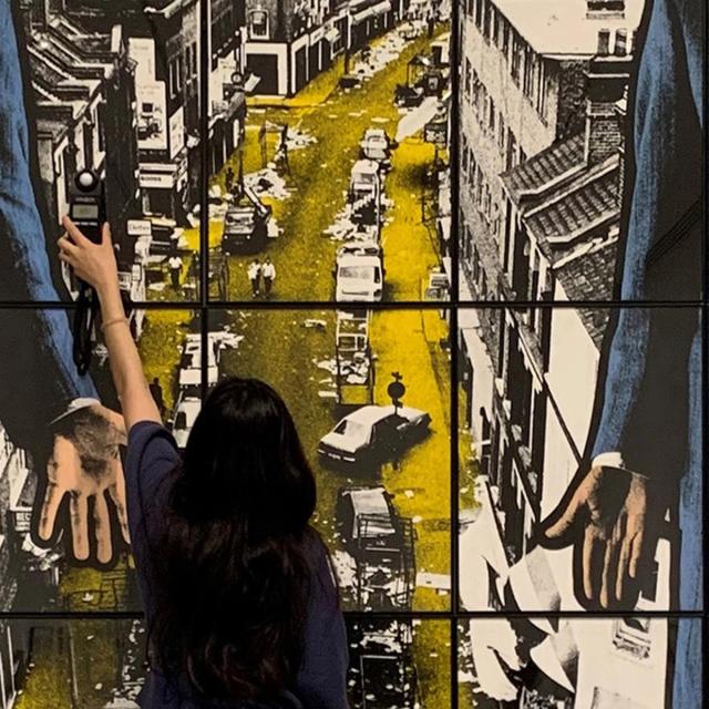 Woman facing large grid of color photographs, reaching up with small light measuring tool