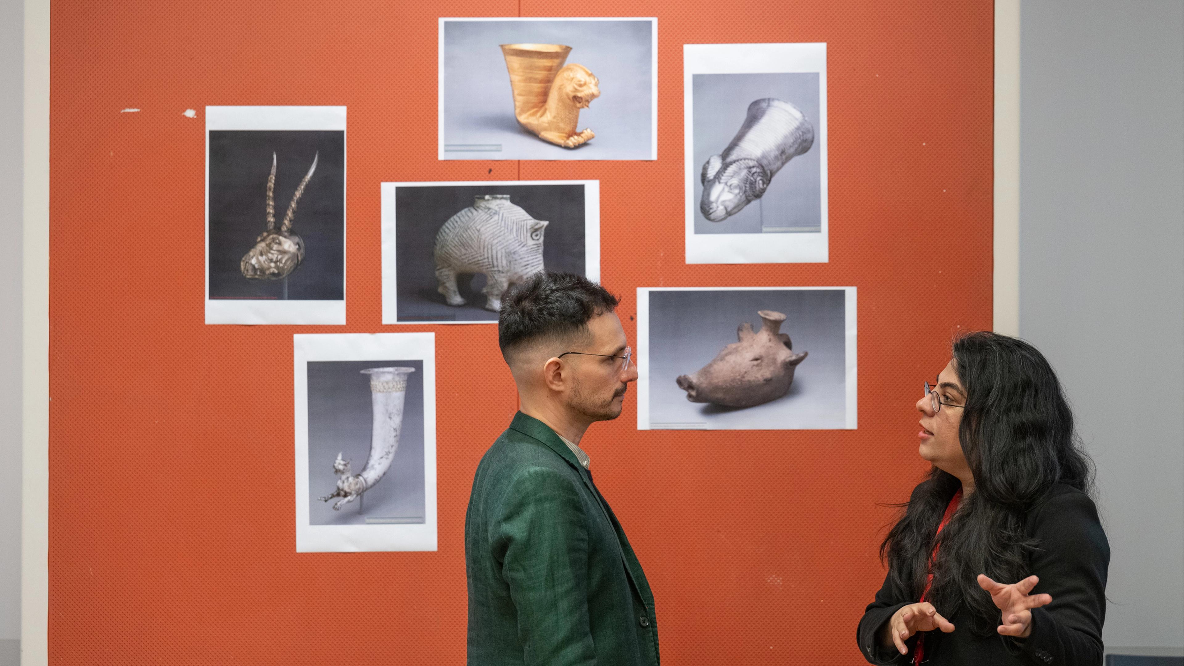 Two people engage in discussion in front of an orange wall displaying various artistic artifacts in black and white photographs.