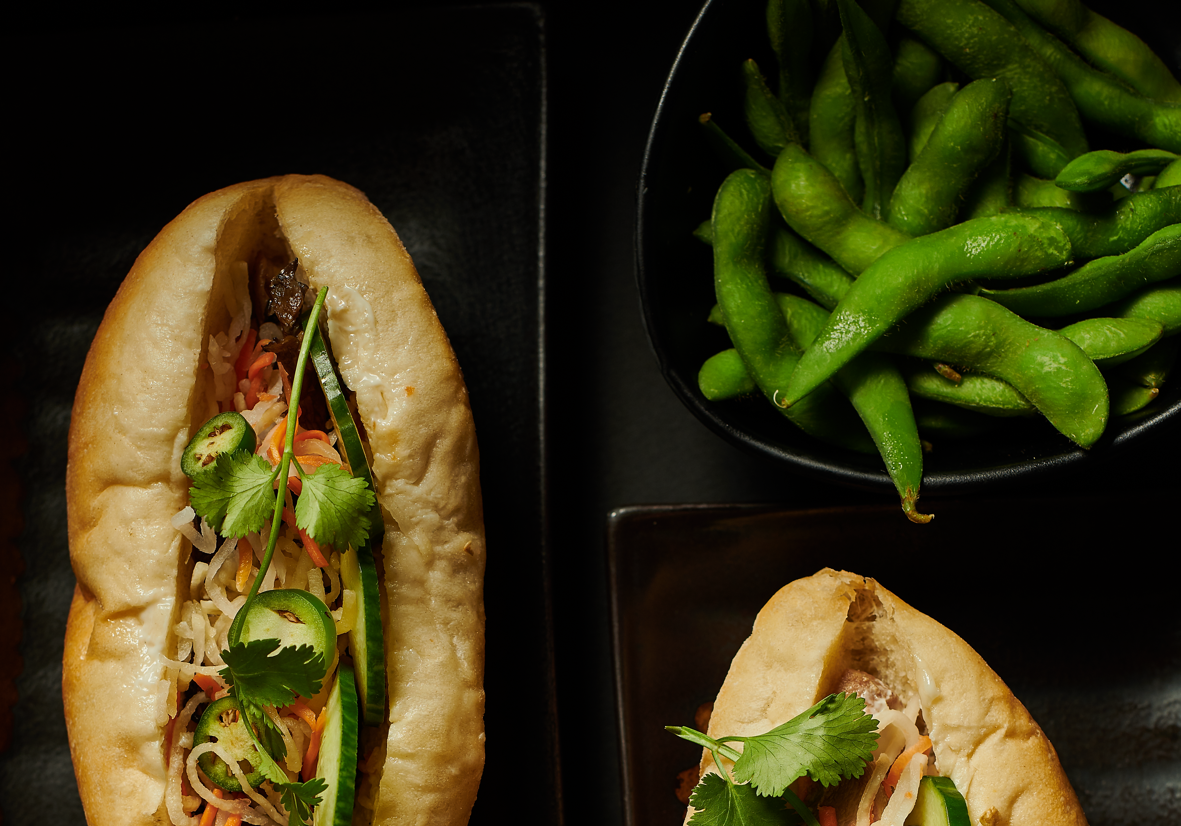 Two bánh mì sandwiches photographed alongside a plate of edamame on a dark background.