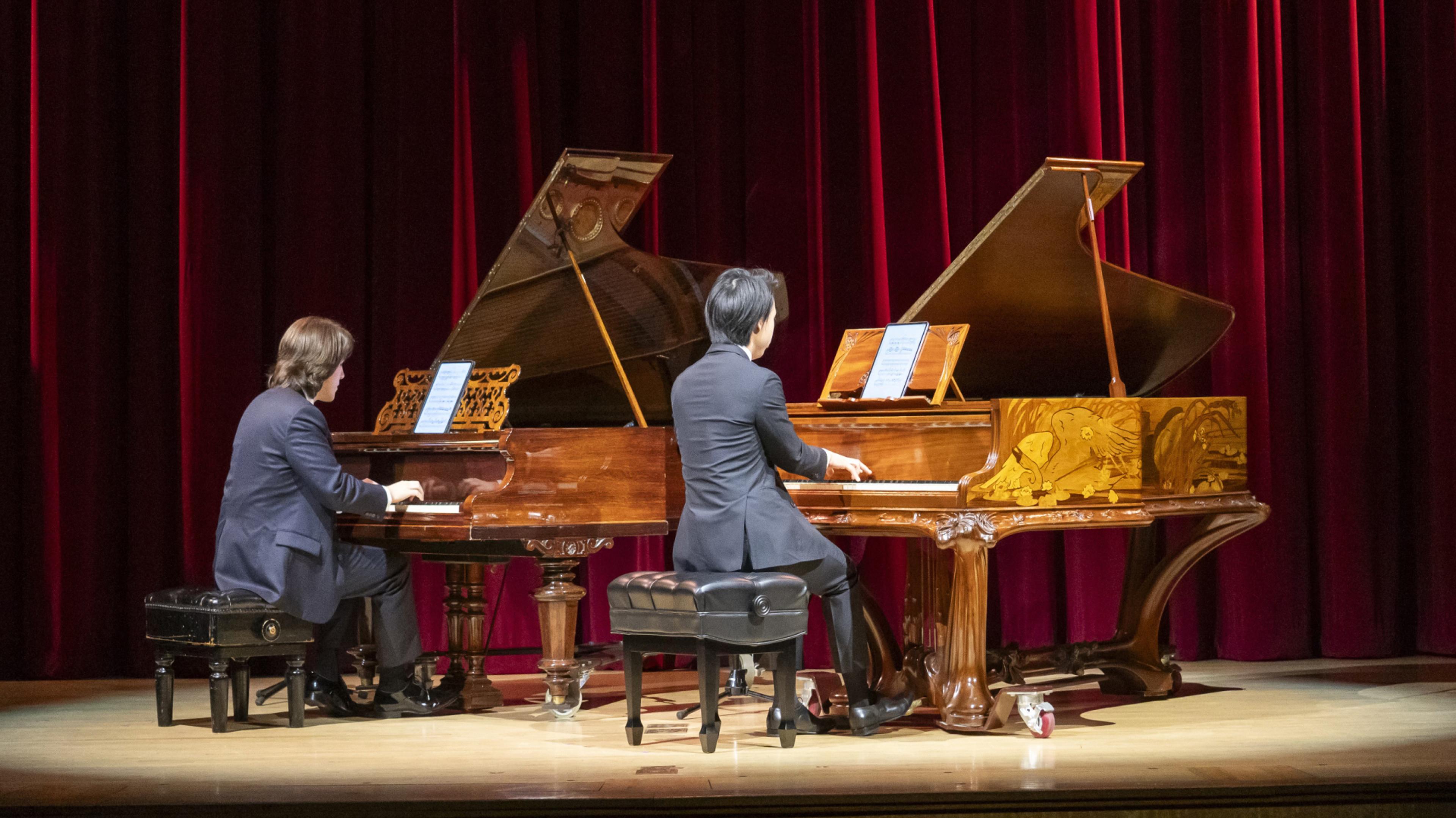 Two men with their backs to the viewer play elaborate grand pianos on a spotlit stage.