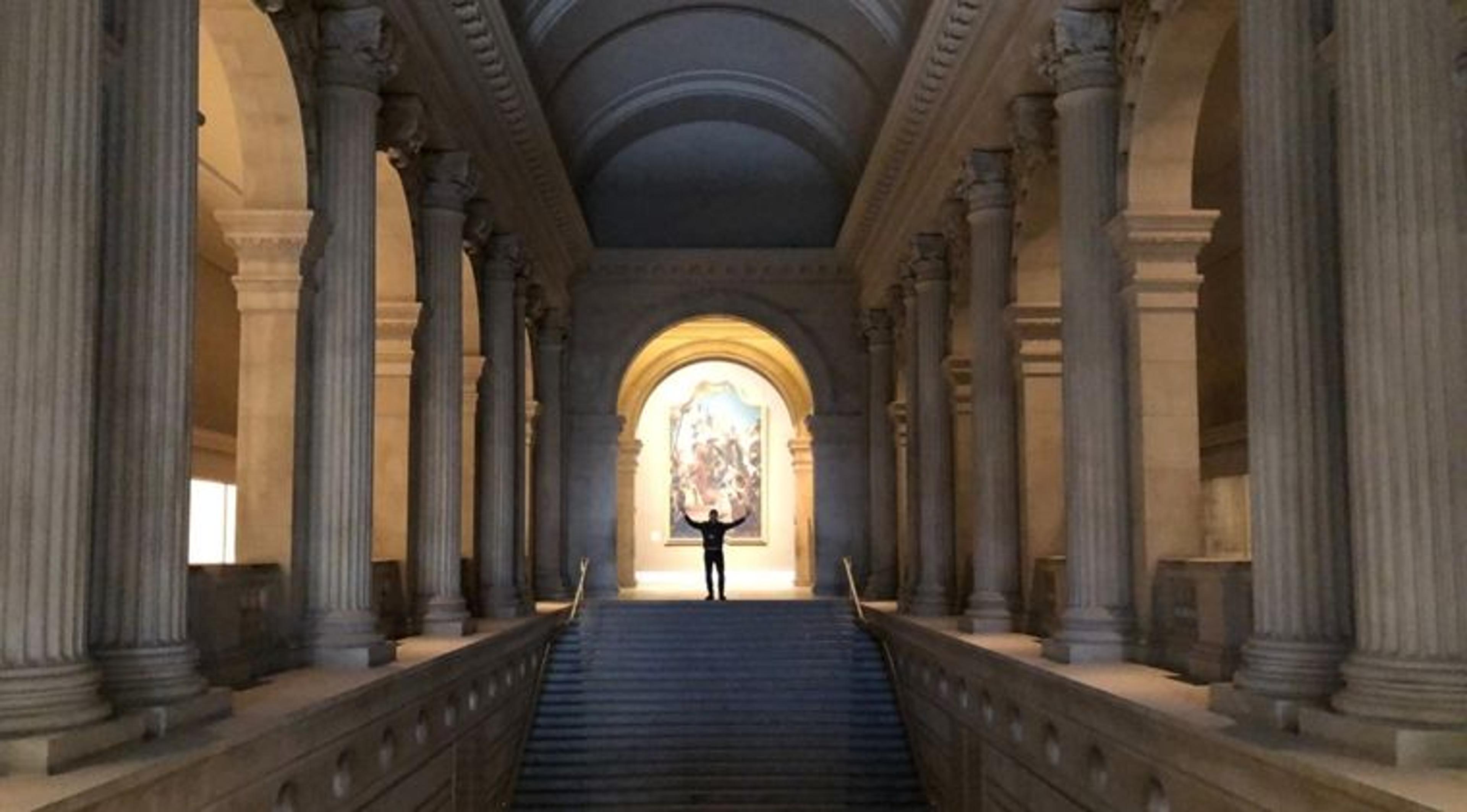 A man stands with his arms outstretched at the top of a large stairwell, surrounded by columns in the Metropolitan Museum of Art