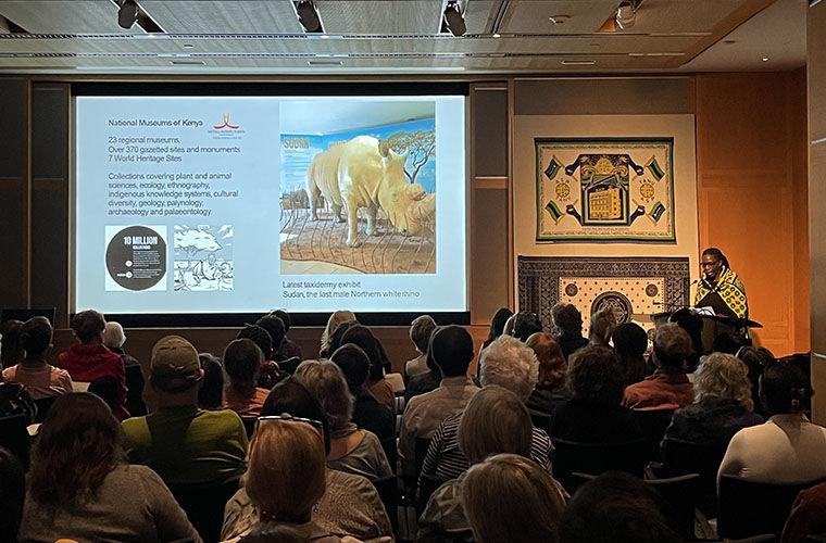 A woman in a yellow patterned shawl speaks in a crowded auditorium with a projection featuring a rhinoceros.