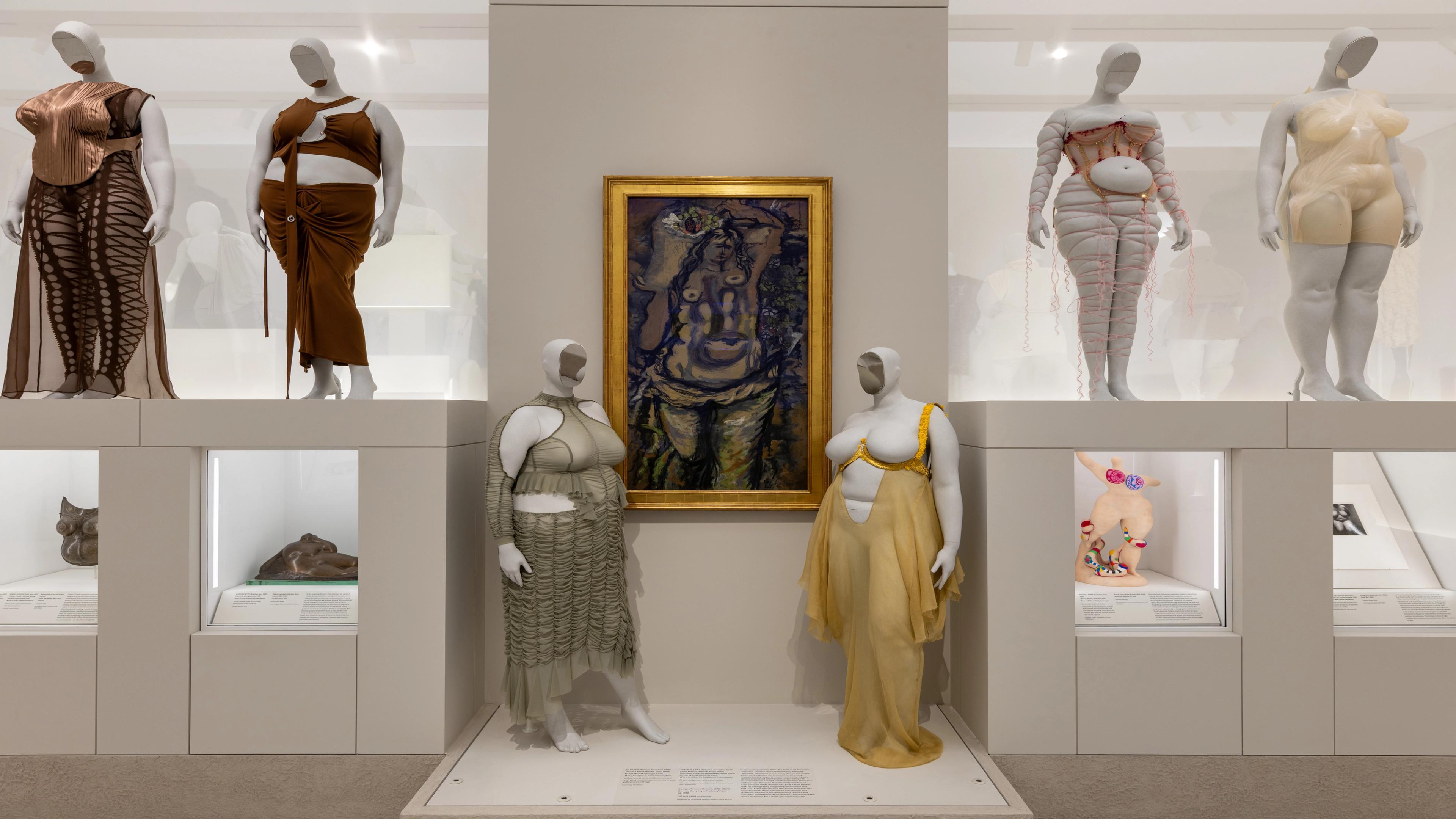 Museum exhibit with mannequins in avant-garde clothing surrounds a central abstract painting in a golden frame. 