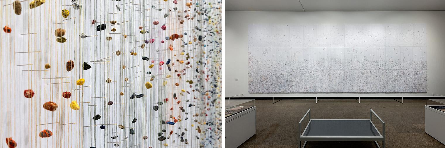 Close-up of colorful stones suspended by lines in an intricate pattern, adjacent to a gallery wall displaying a large, abstract artwork composed of subtle hues.