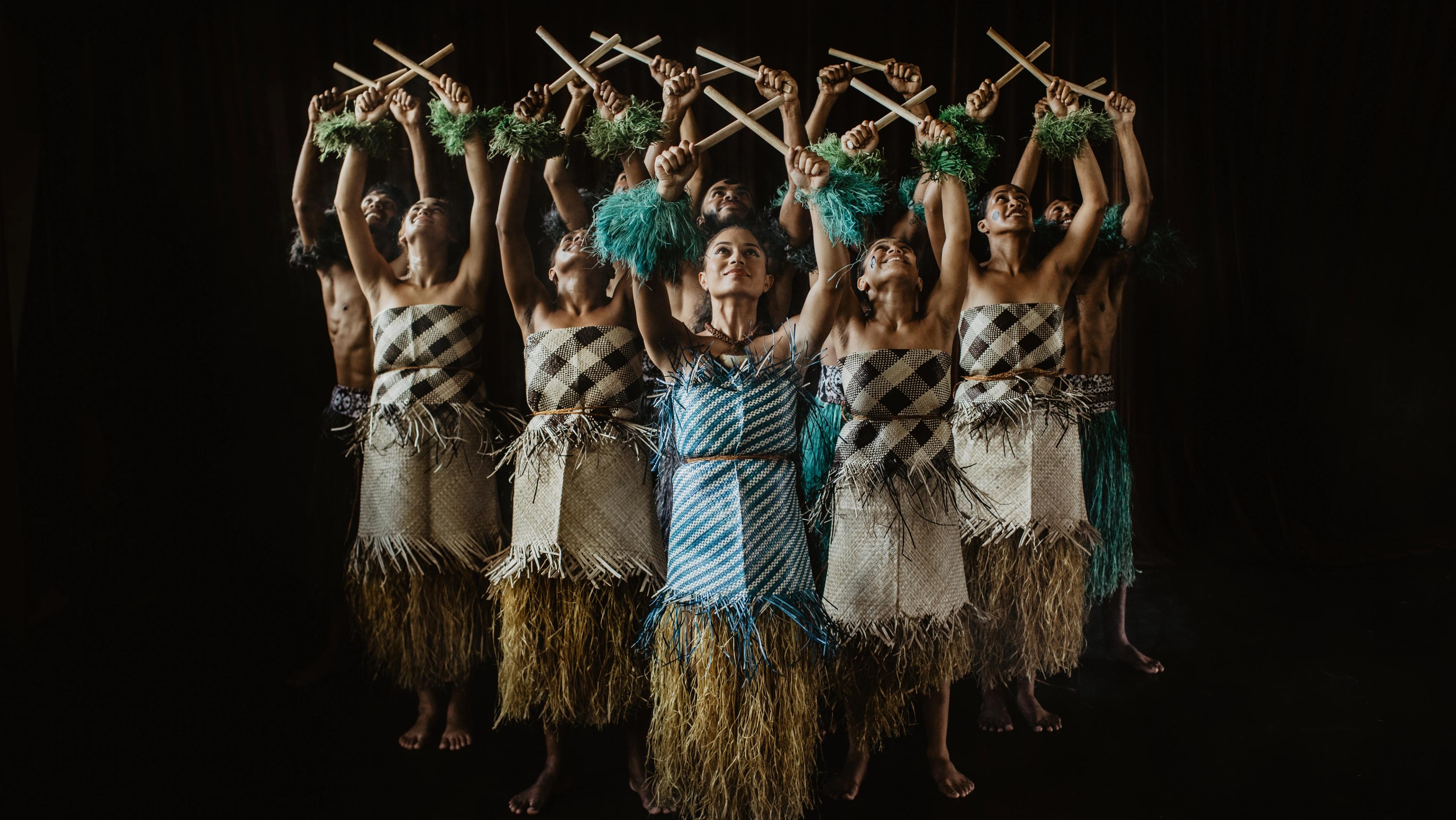 A group of dancers in traditional Fijian clothing hold sticks in an X pattern above their heads.