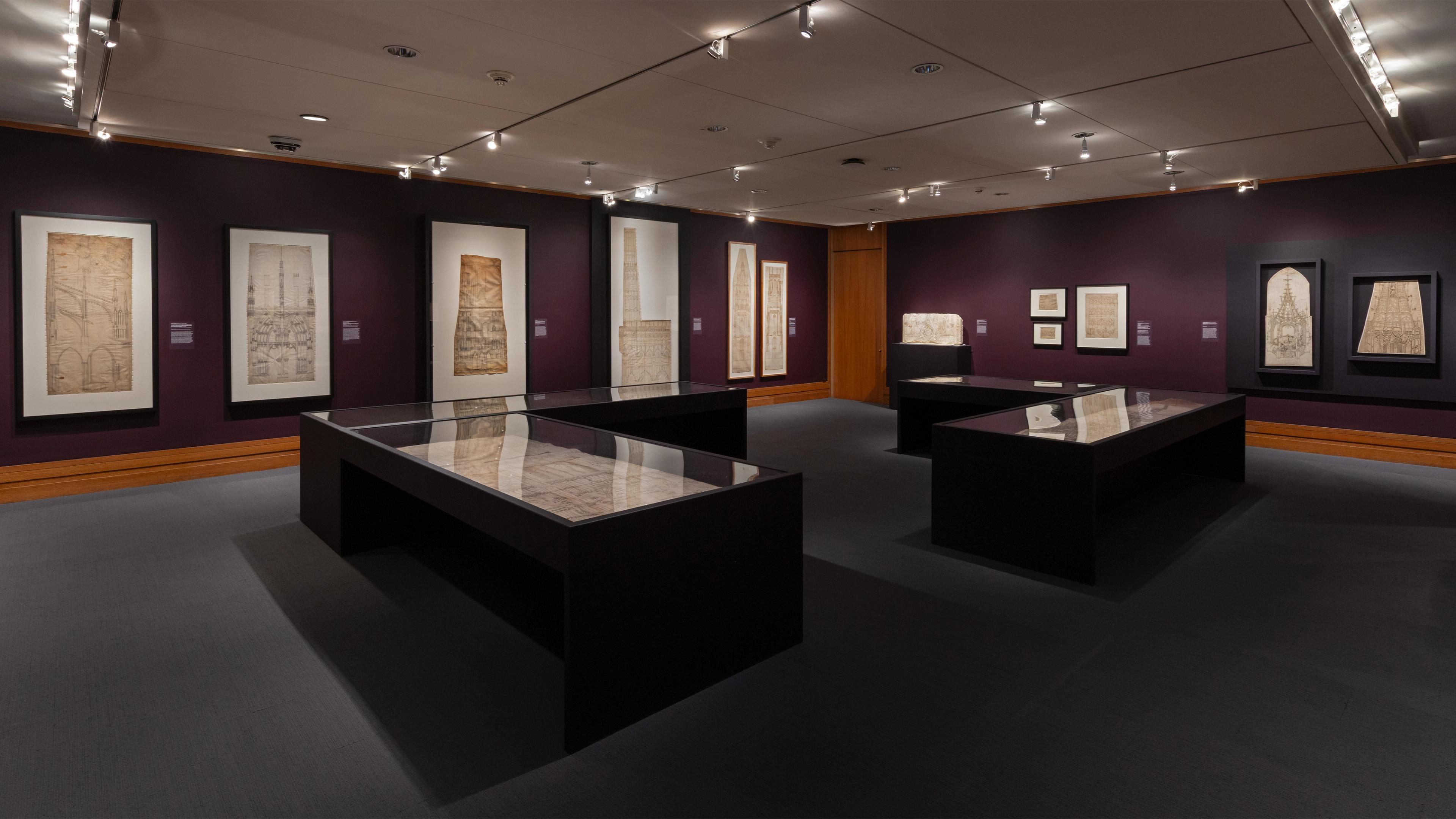 A dimly lit museum gallery with dark walls displays architectural drawings in frames and glass cases.