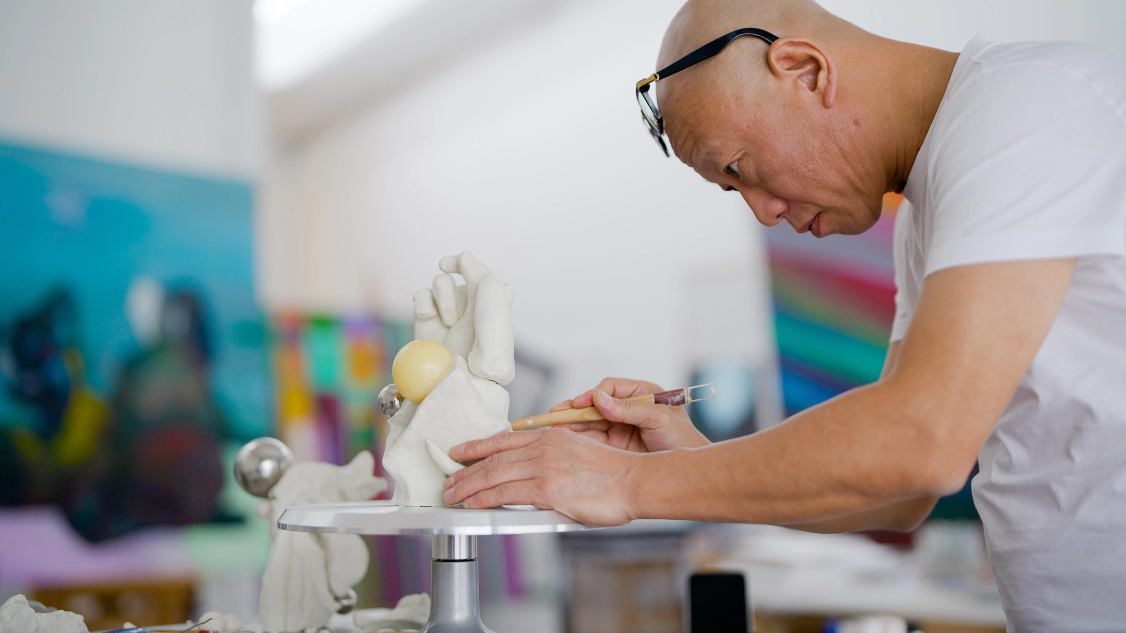 Person leaning over a small pedestal table, shaping a white sculpture with a handheld tool. Colorful artworks are blurred in the background.