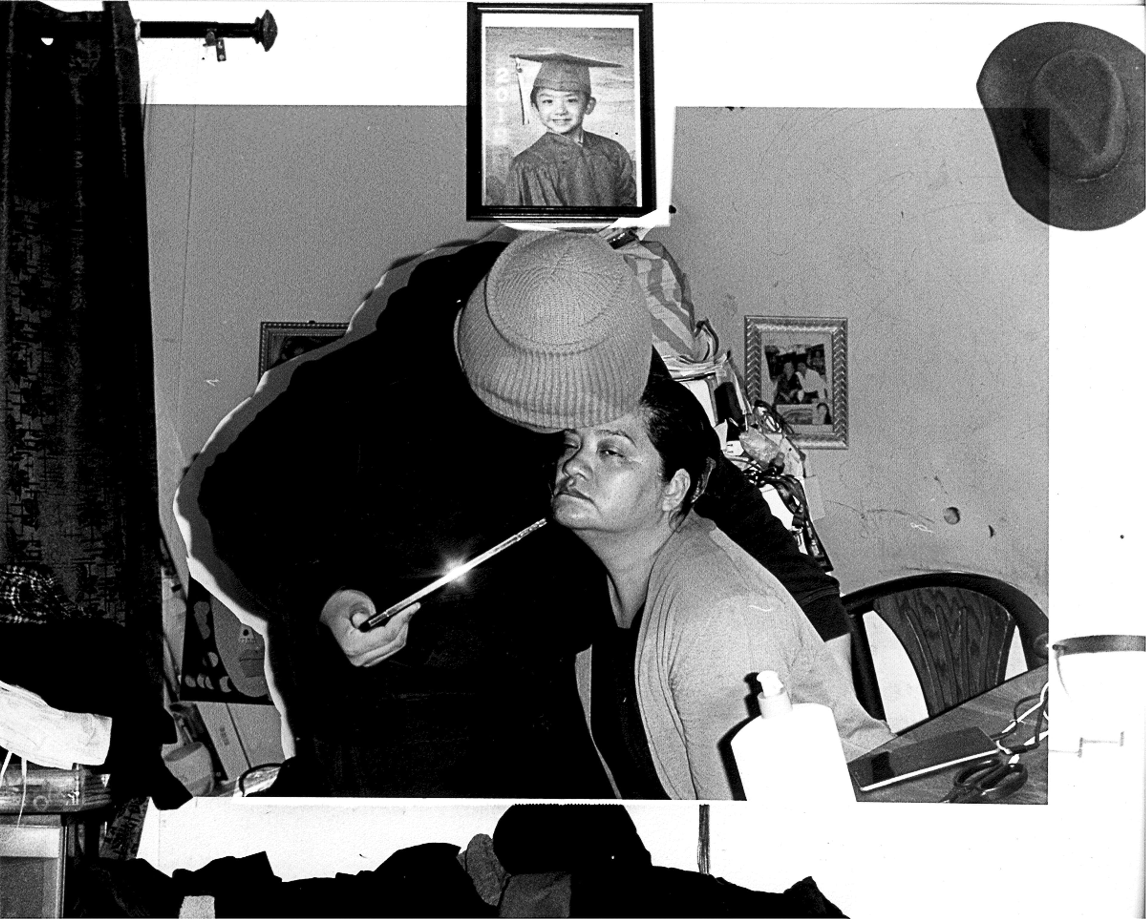 A collage of black and white photographs, with drapes, a hat, and a framed photograph of a child wearing a graduation cap surrounding a central image of a man standing over a seated woman, pointing a reflective object at the woman's chin.