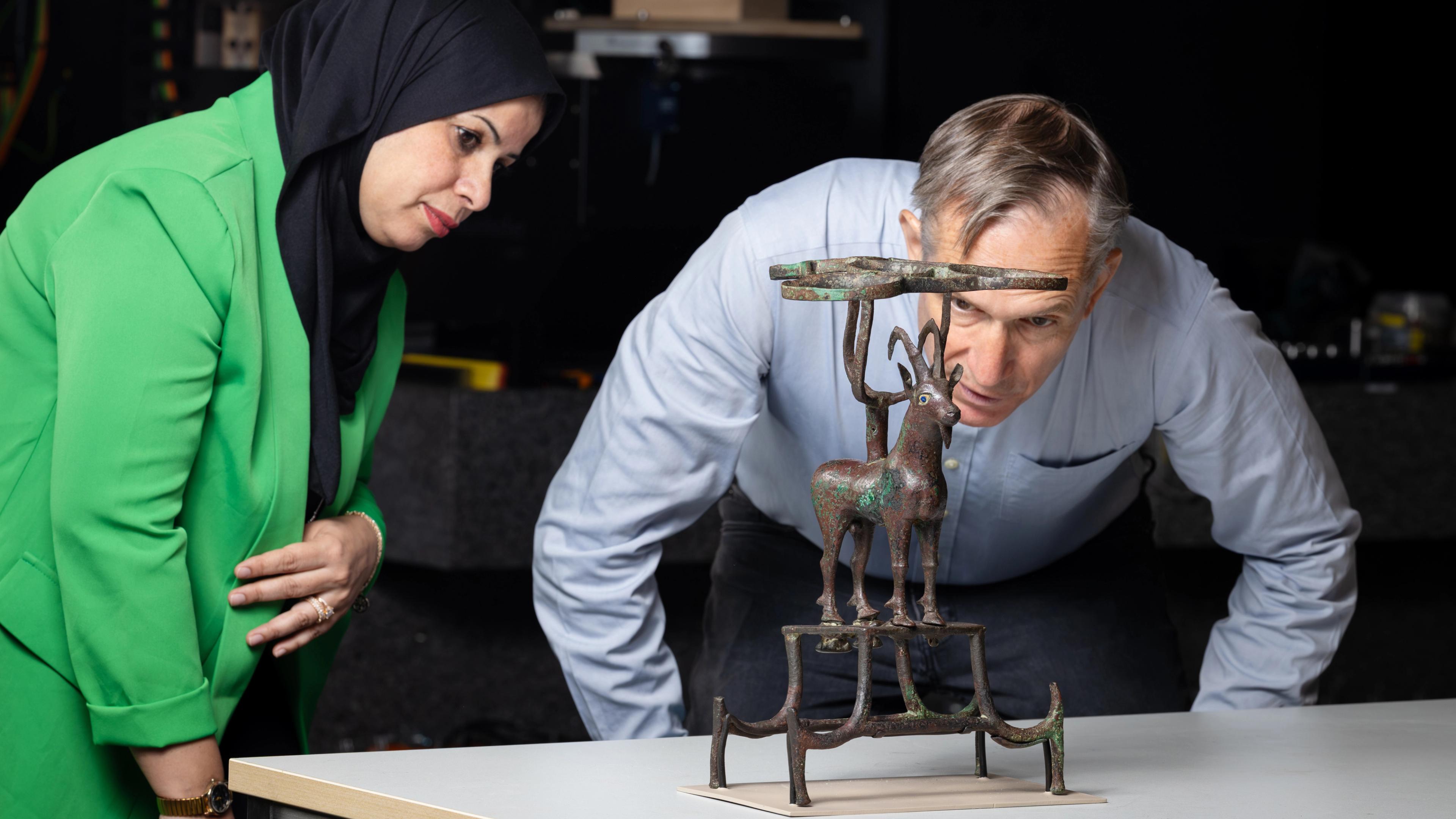 a woman in a green blazer and black headscarf and a man in a blue shirt observing an ibex sculpture