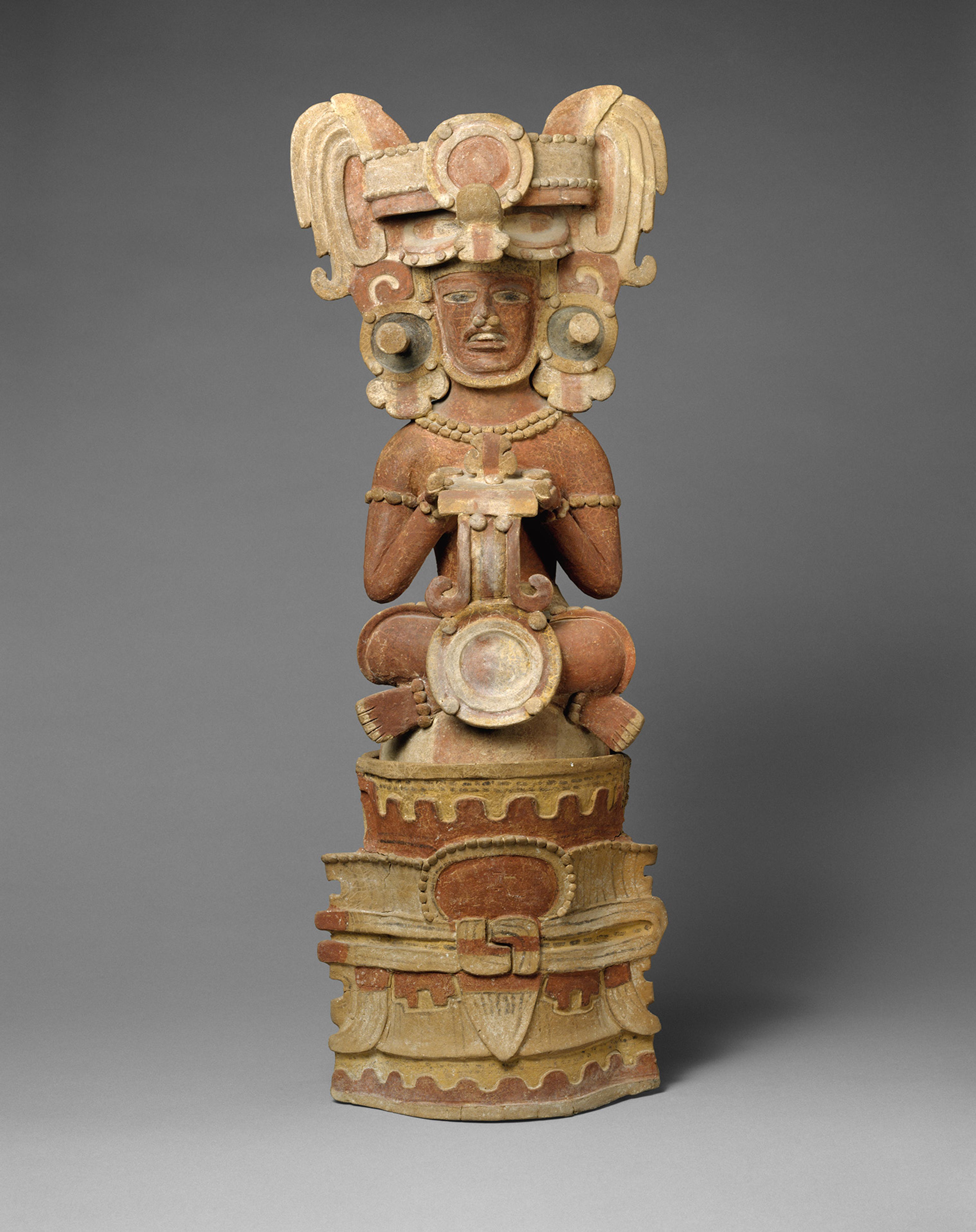 Ornate Mesoamerican-style sculpture of a seated figure with an elaborate headdress and jewelry, resting on a carved pedestal against gray.