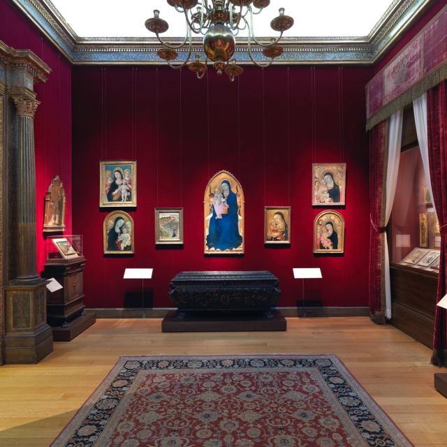 A room within the Robert Lehman Collection at The Met with red walls displays framed Renaissance religious paintings, a dark ornate chest, and wooden furniture.