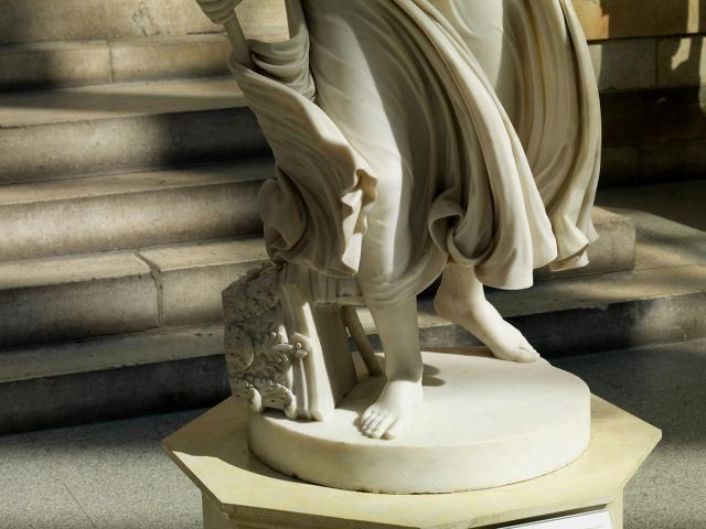 An image of the almost life-sized marble white sculpture Nydia, the blind flower girl of Pompeii, of her lower half and her flowing drapery taken in the American Wing Engelhard Sculpture Court at The Met, a skylit space with direct, dramatic natural light. Her feet are visible at the round base of the sculpture, along with a fallen Corinthian column and her staff along her right food. There are steps in the background of the shot.