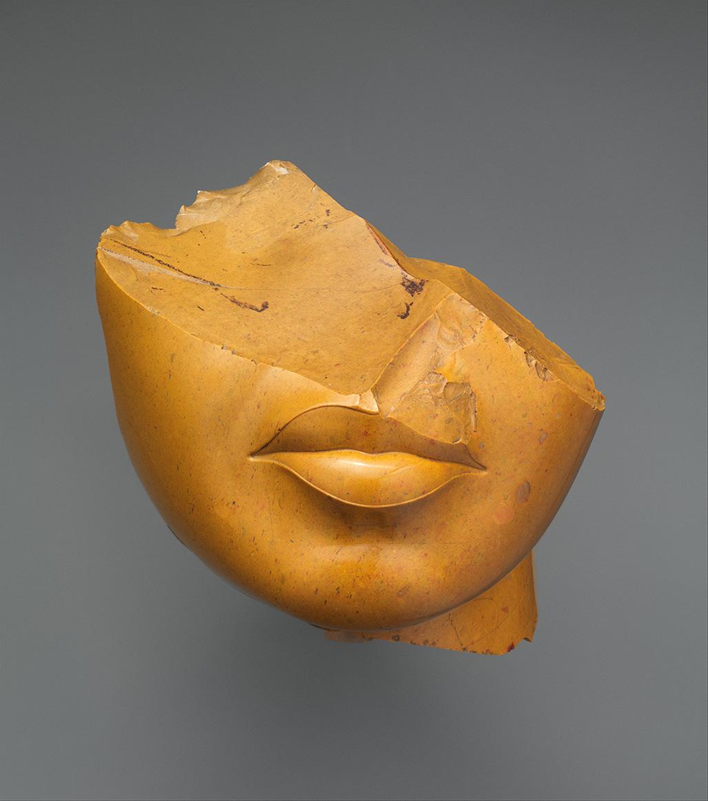 A fragment of a yellow-brown stone sculpture showing the lips and chin of a human face, with the upper portion broken away.