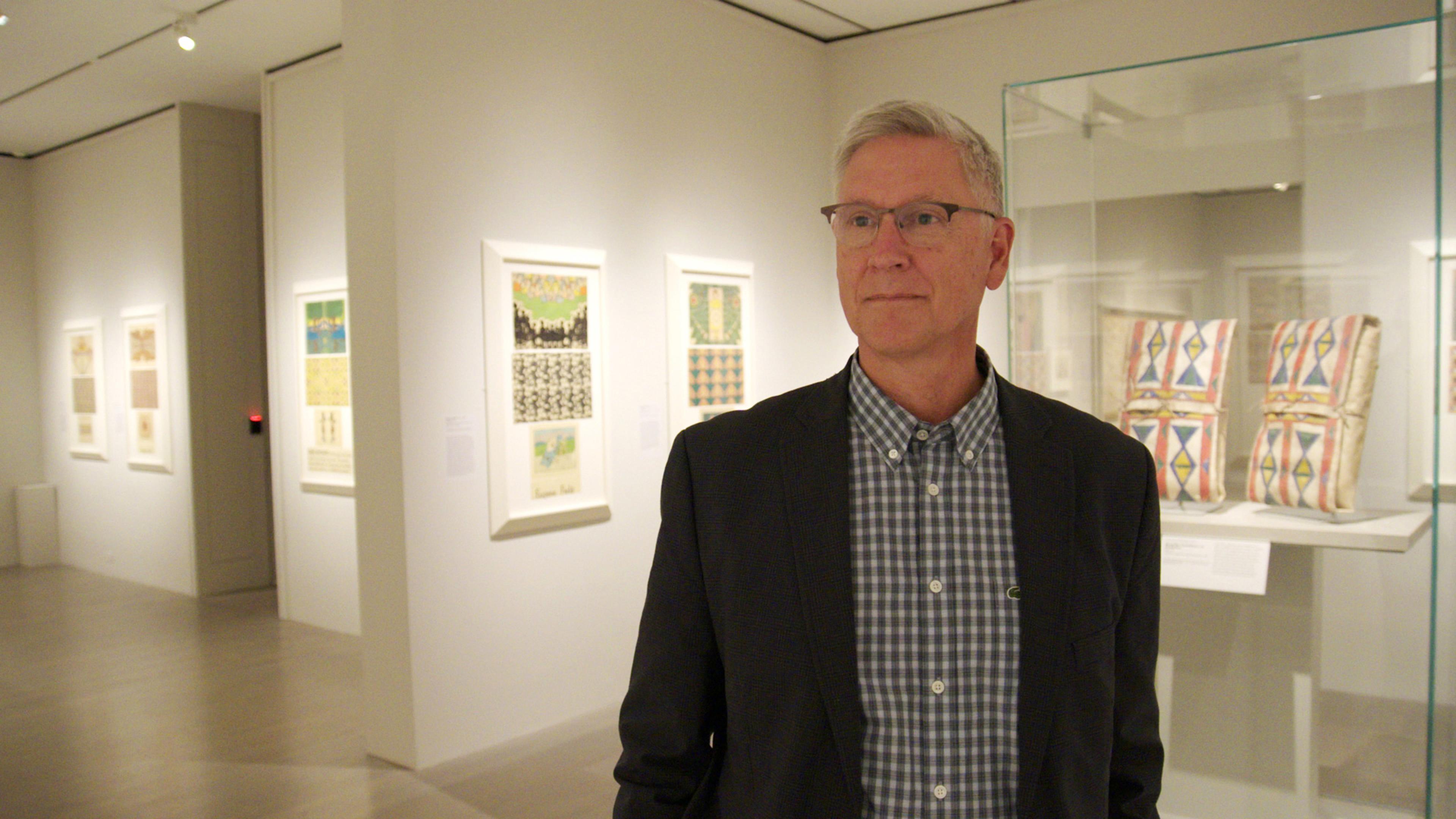 Philip Deloria, in a dark suit jacket and patterned shirt, stands in a gallery in front of various prints and objects with geometric patterns