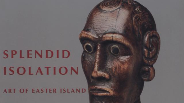 Close-up of a dark wooden sculpture of a head with large eyes, intricate ear details, and a solemn expression. Text reads "Splendid Isolation: Art of Easter Island."