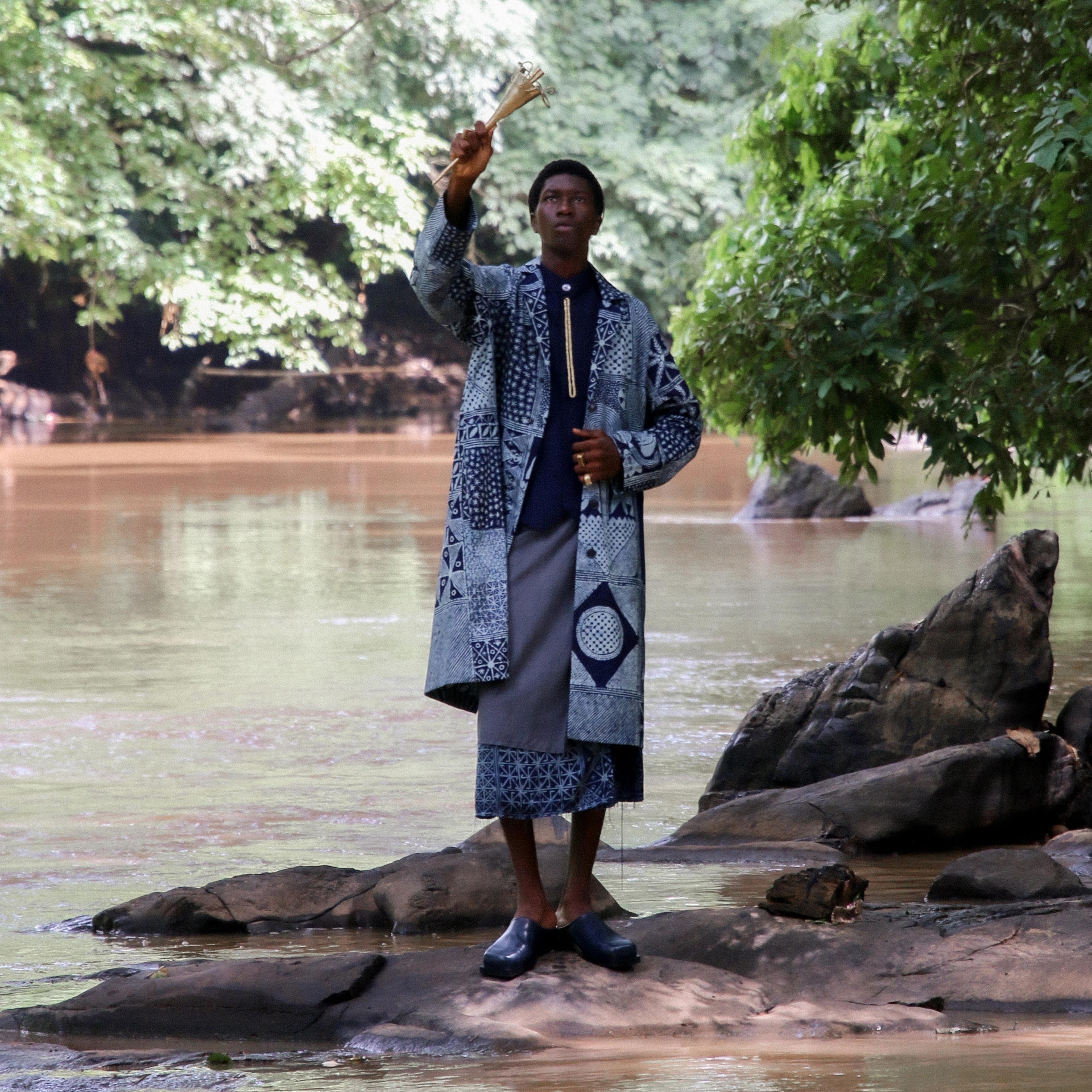 Young Black man standing in a body of water, upright looking up at the sky. He wears traditional African attire, while one hand extends above him.