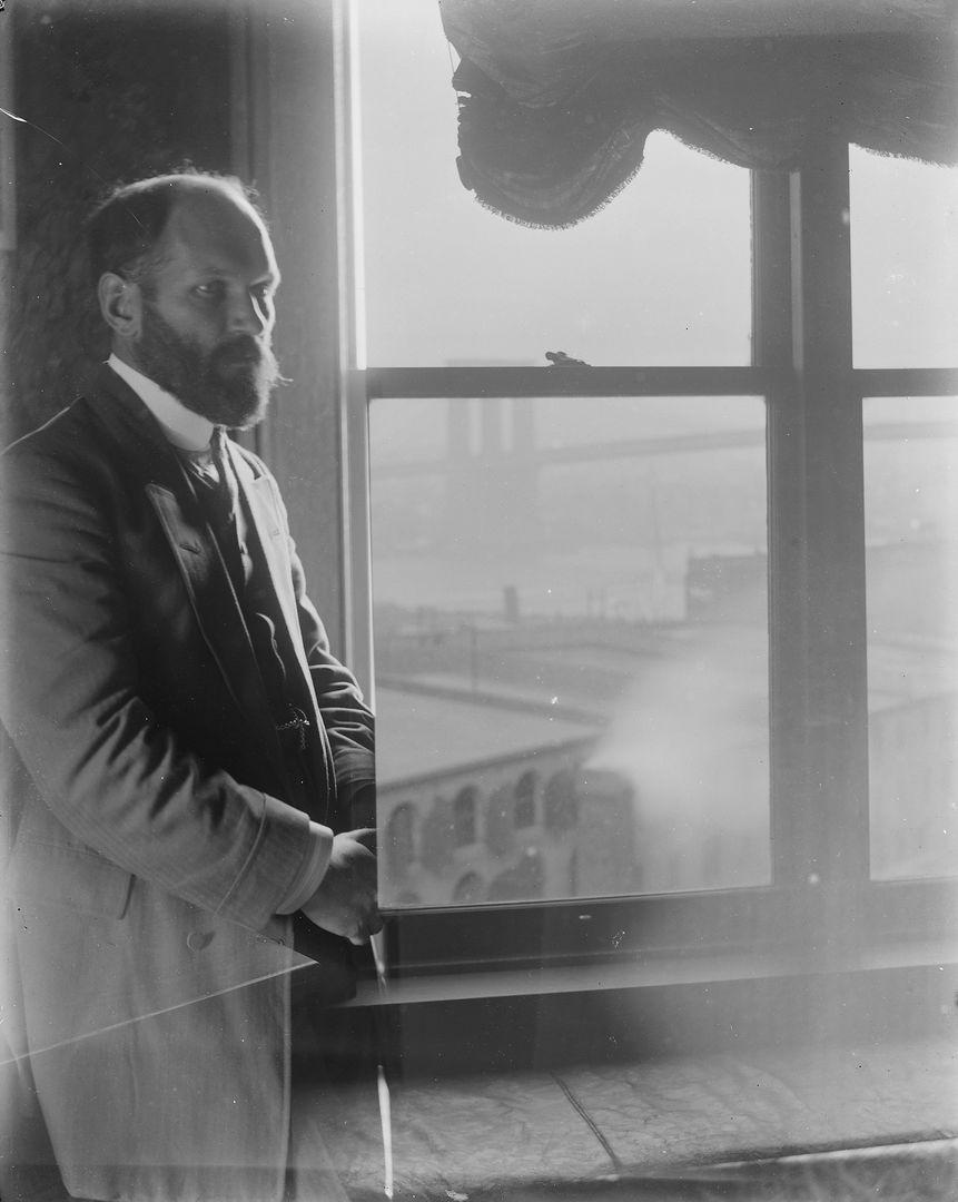 Black and white Image of a bald white man in a suit with a beard. He is standing to the left of a large window.