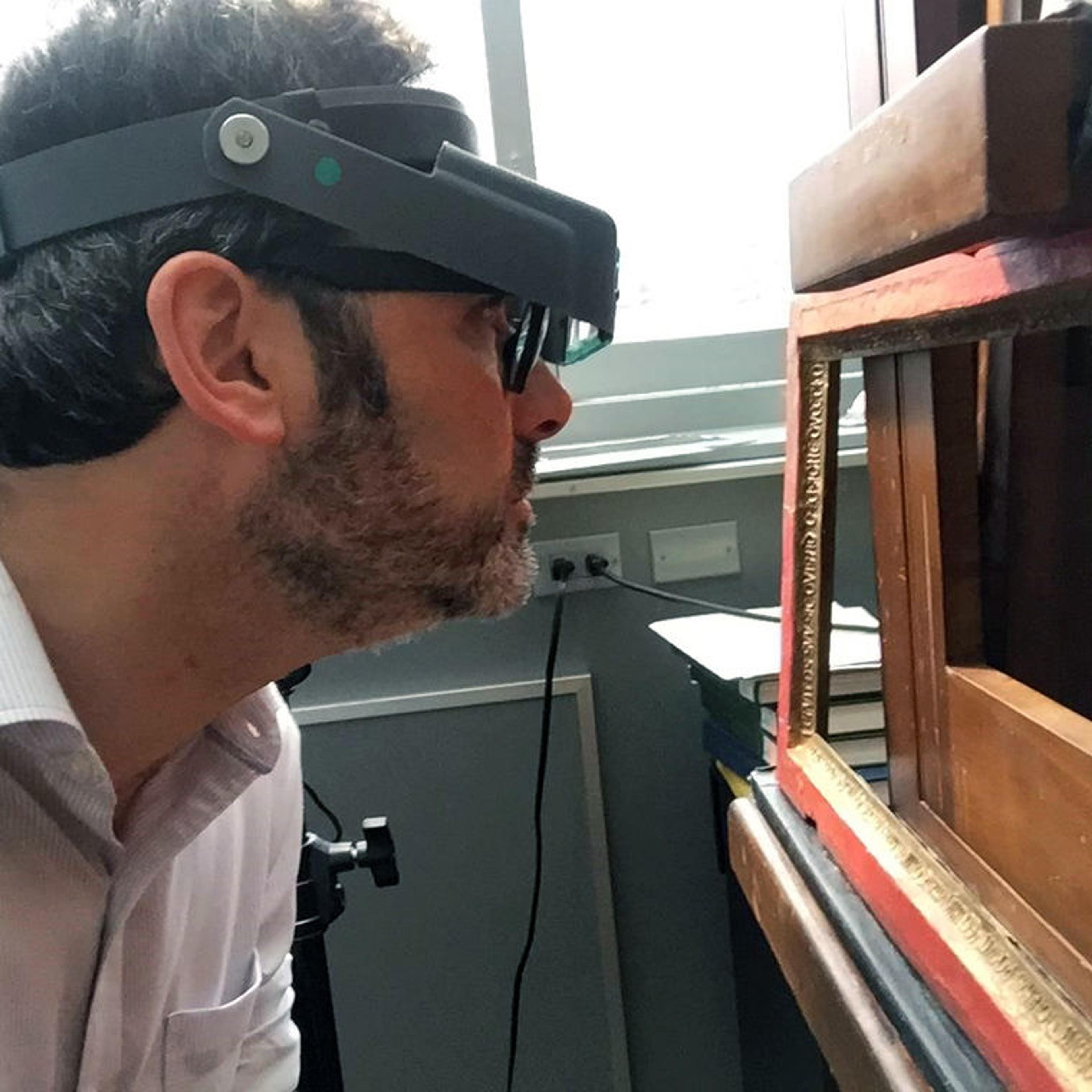 A man wearing a headpiece with magnifying glass closely inspects a wooden painting frame in a sunlit conservation studio