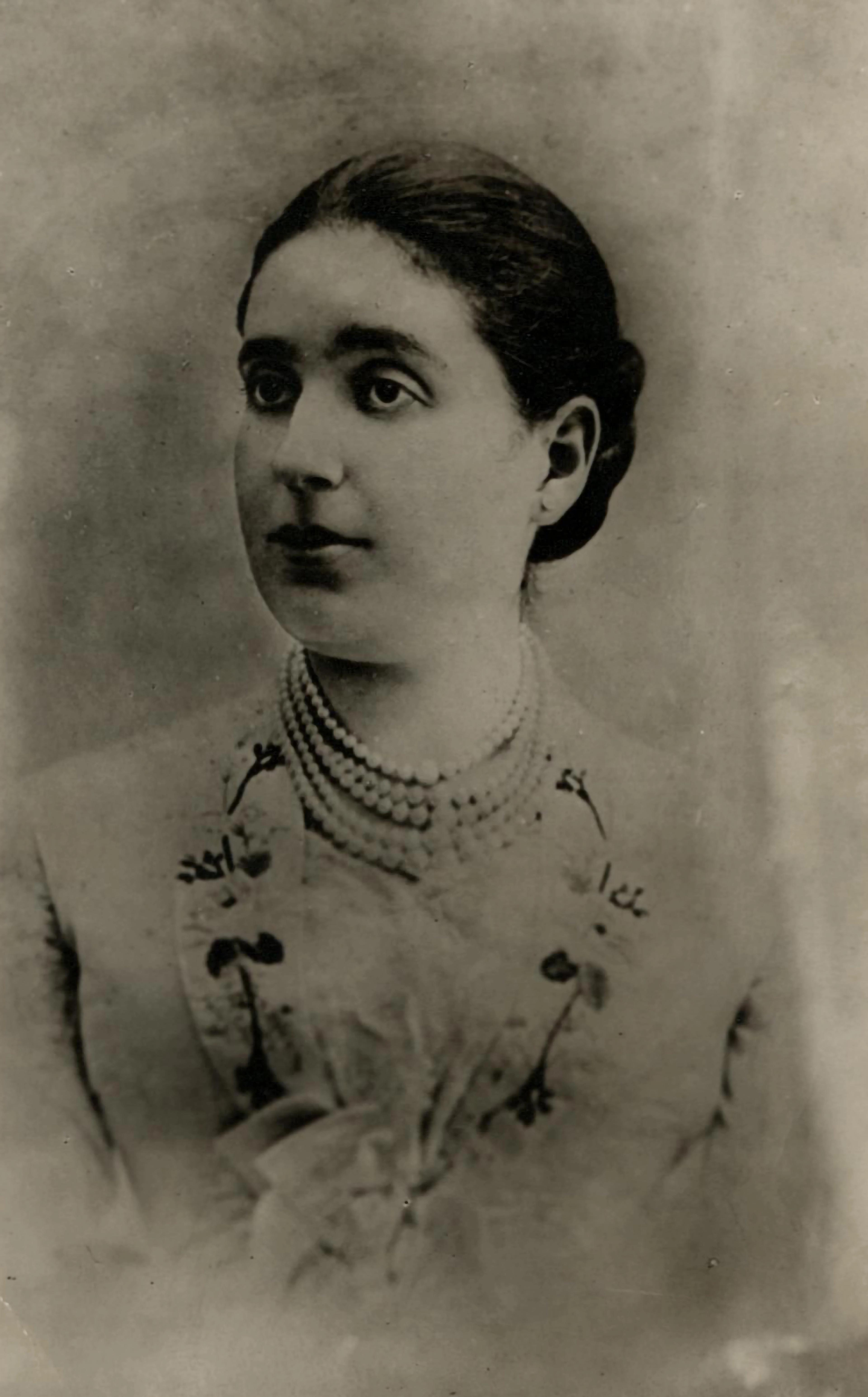 A vintage studio portrait of a woman wearing a high‑neck garment and layered pearl necklaces. Her hair is styled neatly and pulled back.