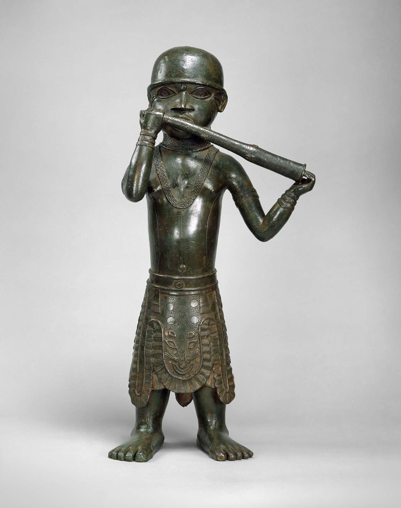 Bronze statue of a standing figure wearing a detailed skirt and playing a long flute-like instrument, with stylized features and intricate jewelry.