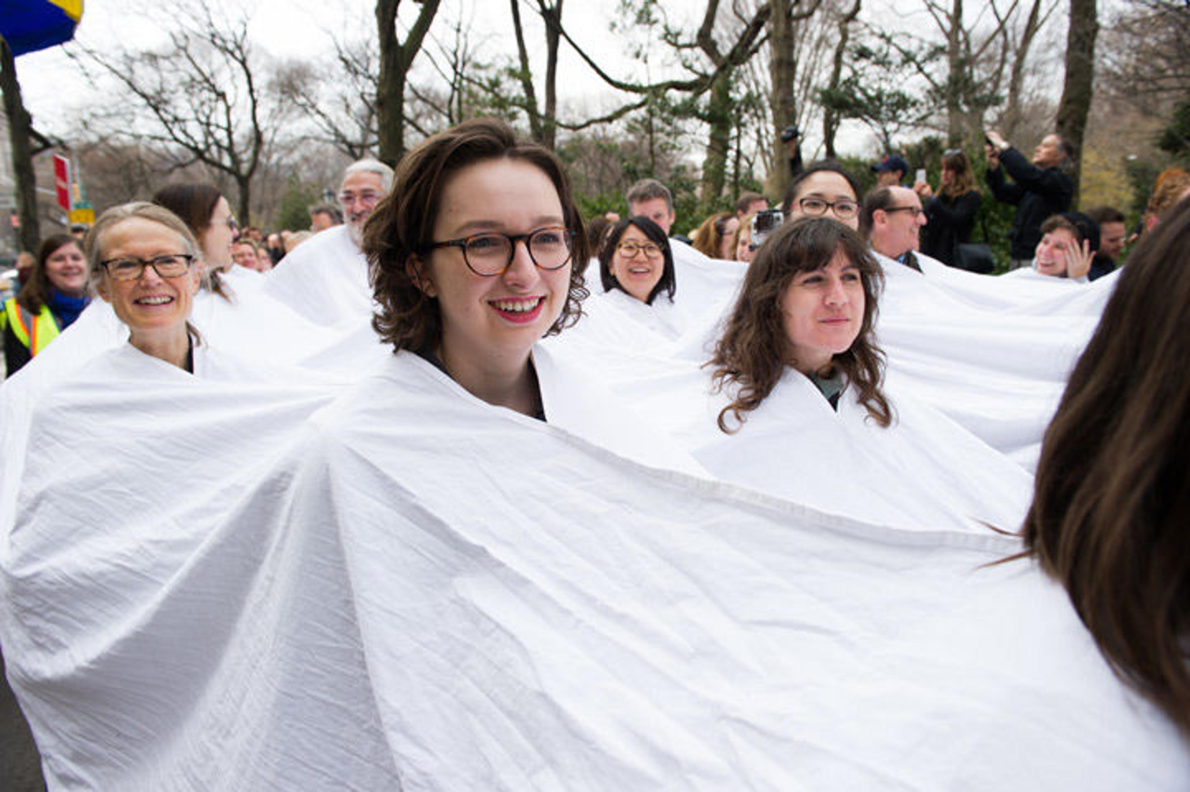 A group of people take part in a procession north on Madison Avenue while entrenched in a large piece of white cloth from which only their heads emerge