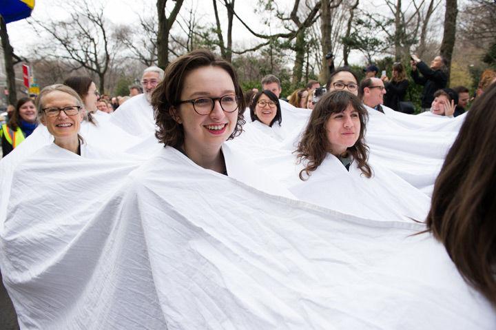 A group of people take part in a procession north on Madison Avenue while entrenched in a large piece of white cloth from which only their heads emerge