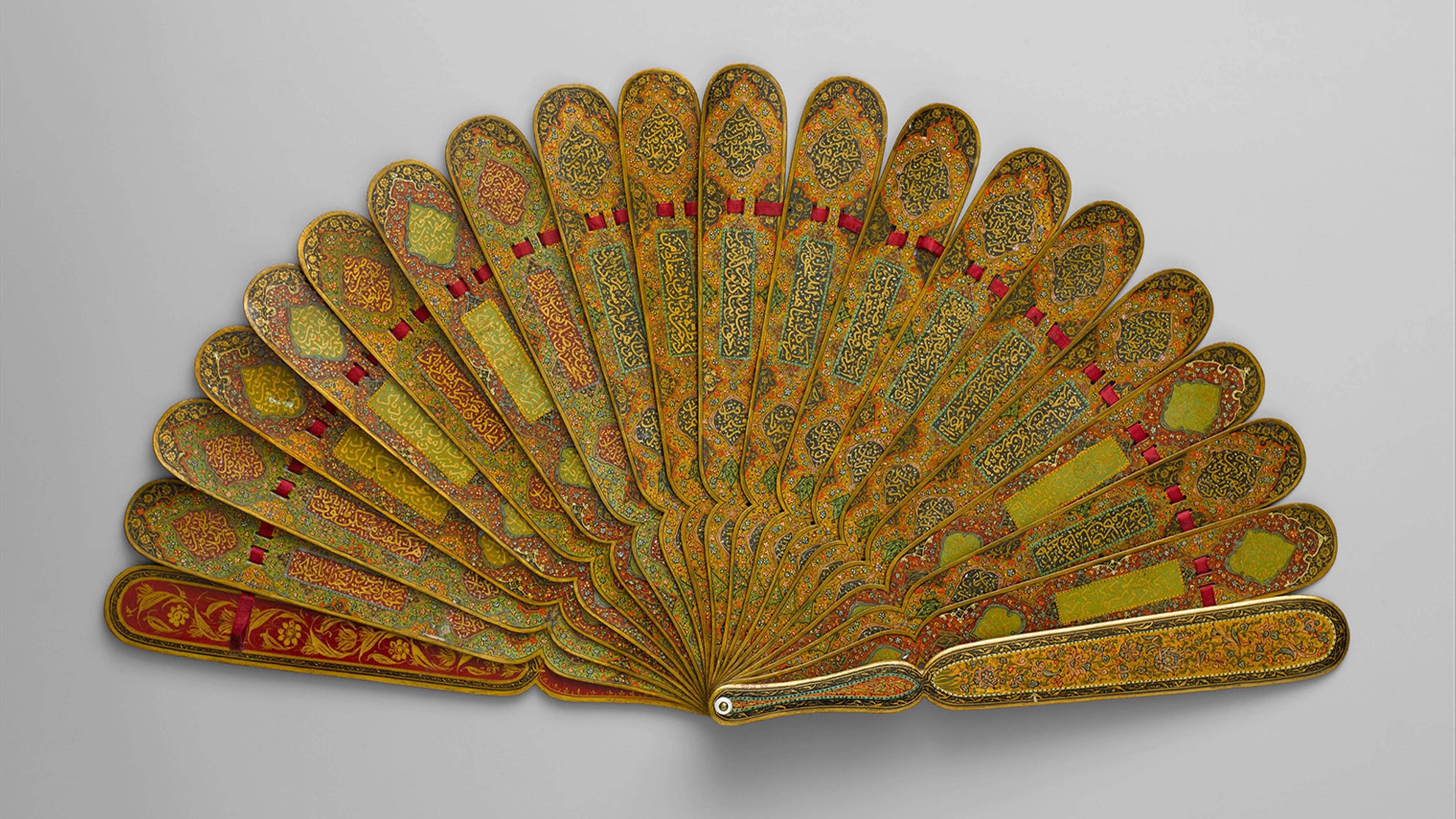 Ornate folding fan with golden patterns and red accents on each rib, intricately designed with floral motifs, displayed against a neutral background.