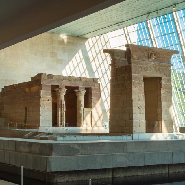 An ancient Egyptian temple structure displayed inside a museum, surrounded by a water with sunlight shining through a wall of glass windows.