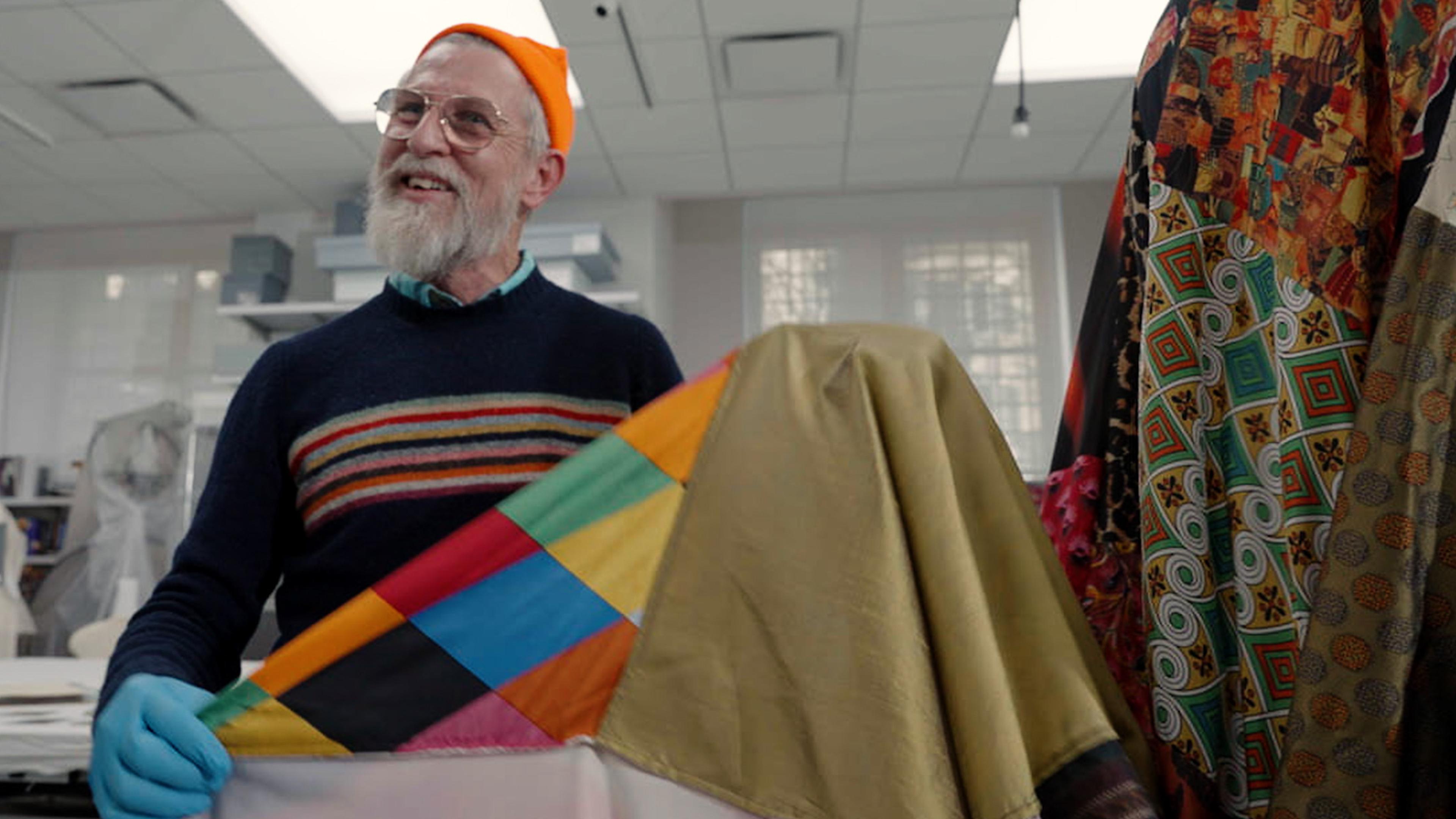 A bearded man wearing a sweater with stripes, glasses, and an orange beanie smiles while holding up a fabric with a multicolored diamond pattern.