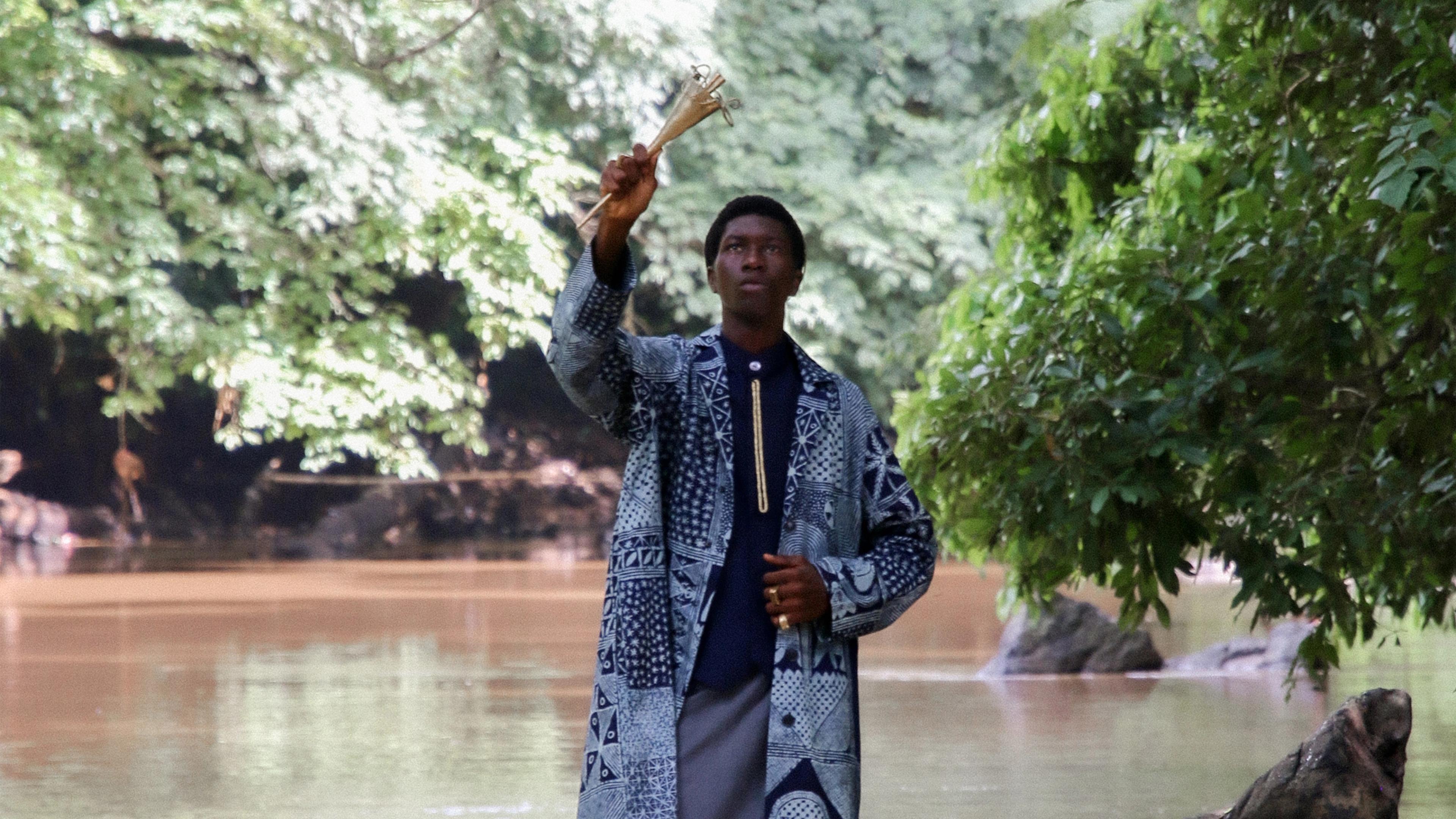 Young Black man standing in a body of water, upright looking up at the sky. He wears a traditional African attire, while one hand extends above him.